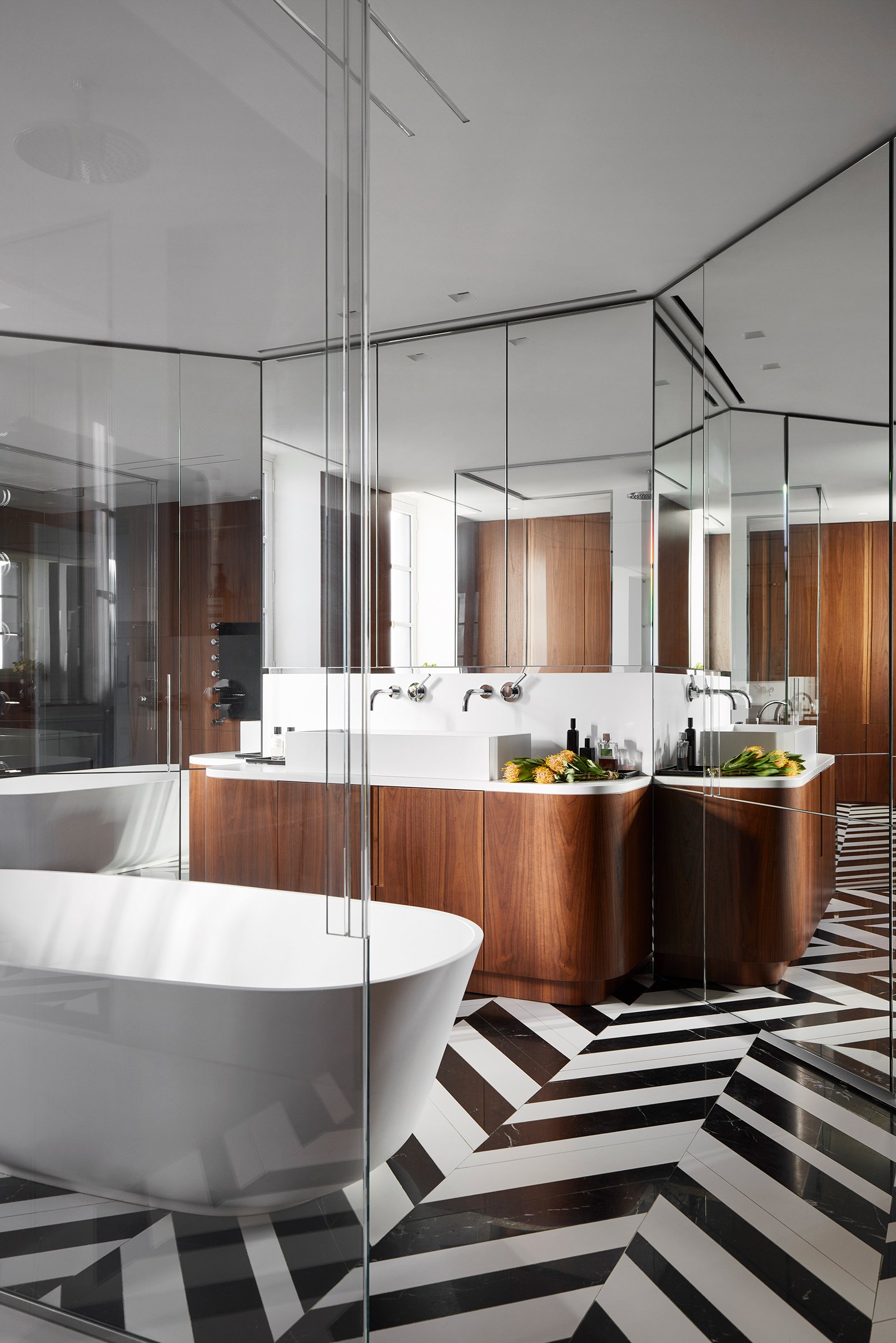 Salle de bain moderne avec baignoire blanche, miroir mural, vanité en bois avec deux lavabos, et un sol à motifs en noir et blanc.