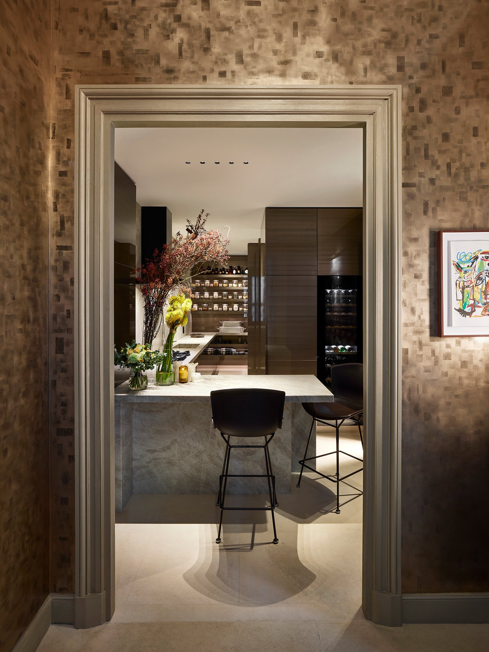 Intérieur d'une cuisine moderne avec un comptoir en marbre, deux chaises noires et une grande vase avec des fleurs, vue à travers une porte encadrée en bois.