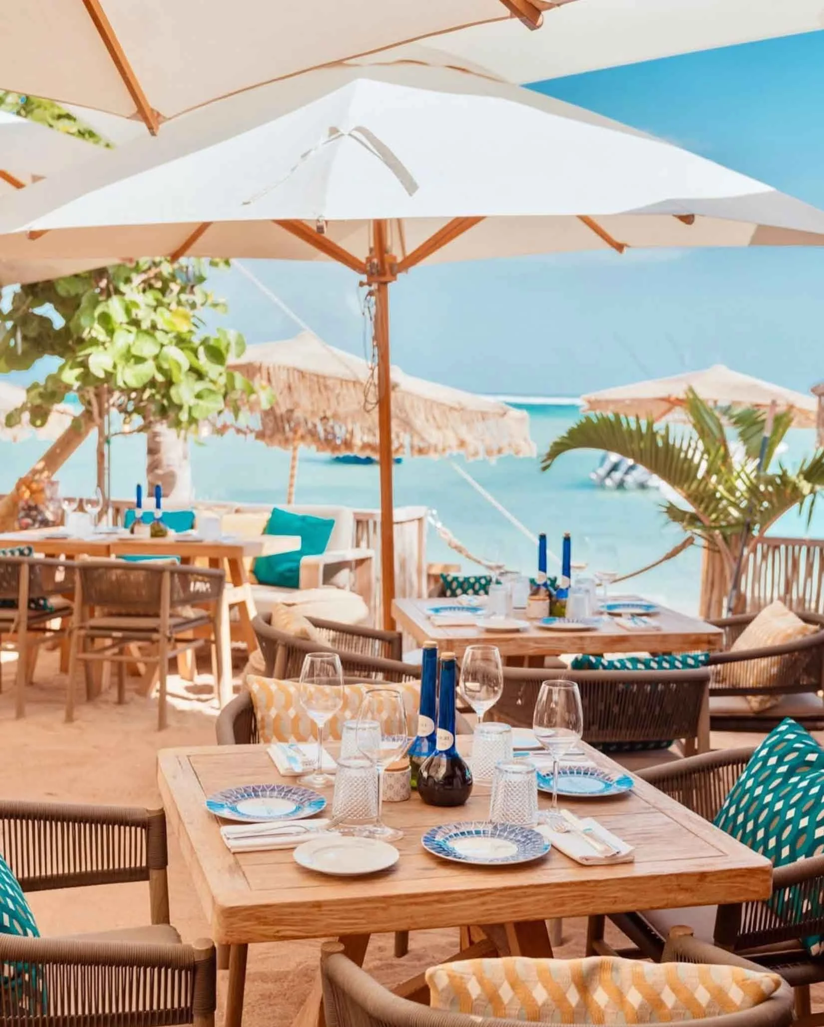 Une terrasse de restaurant en front de mer avec des tables en bois, des chaises et des parasols blancs sous un ciel bleu, face à la mer.