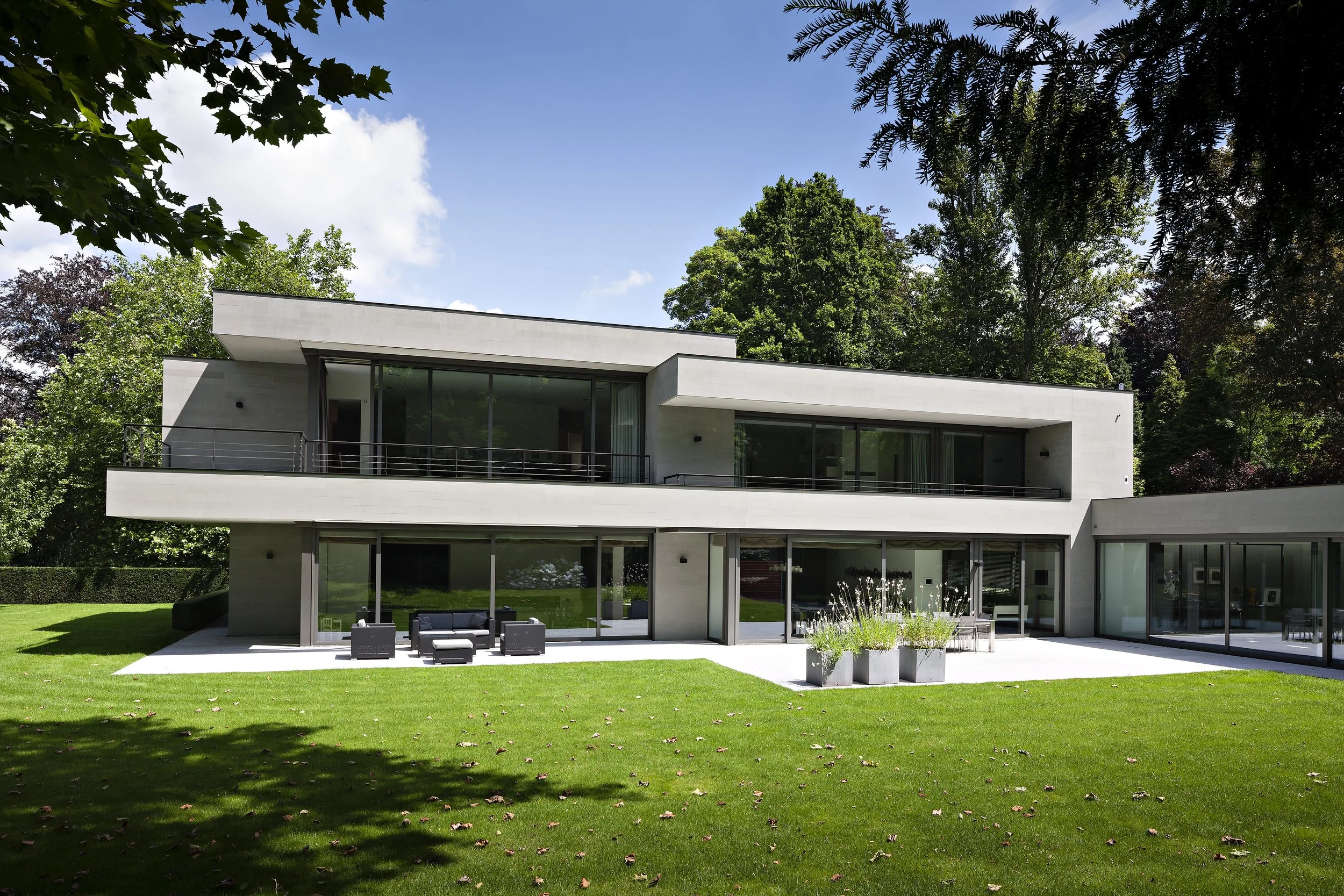 Maison moderne avec grandes fenêtres en verre, terrasse extérieure avec mobilier, sur un terrain vert entouré d'arbres, sous un ciel bleu avec quelques nuages.