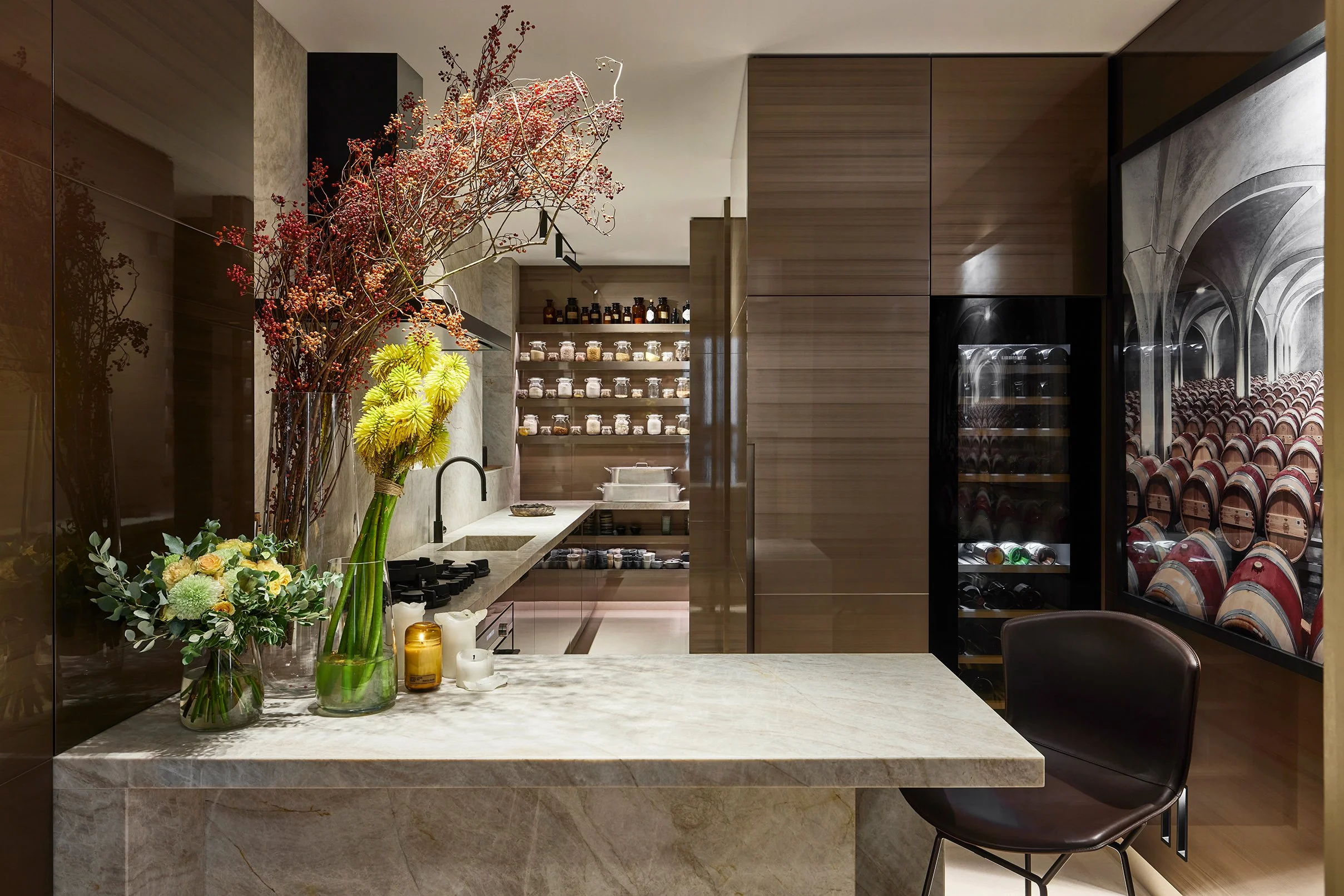 Cuisine moderne avec plan de travail en marbre, étagères de stockage, bouteilles de vin dans un réfrigérateur, et une cave à vin visible à travers une fenêtre. Décor floral avec des fleurs colorées.