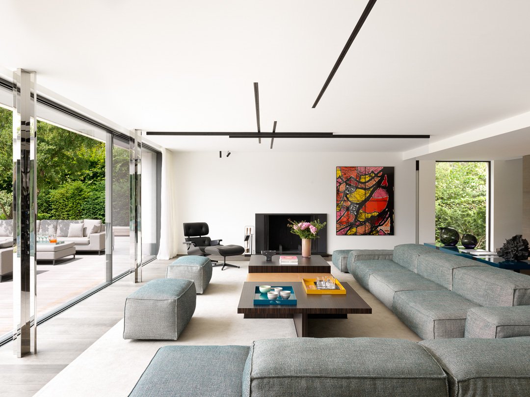 Salon moderne avec grands vitres, canapé gris, table basse, fauteuil noir, tableau abstrait, et vue sur un patio avec mobilier extérieur