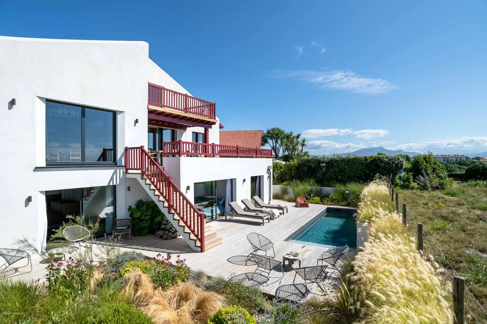 Une maison moderne blanche avec des terrasses en bois rouge, un jardin avec des chaises longues, une piscine, un espace extérieur avec des plantes et un ciel bleu clair.