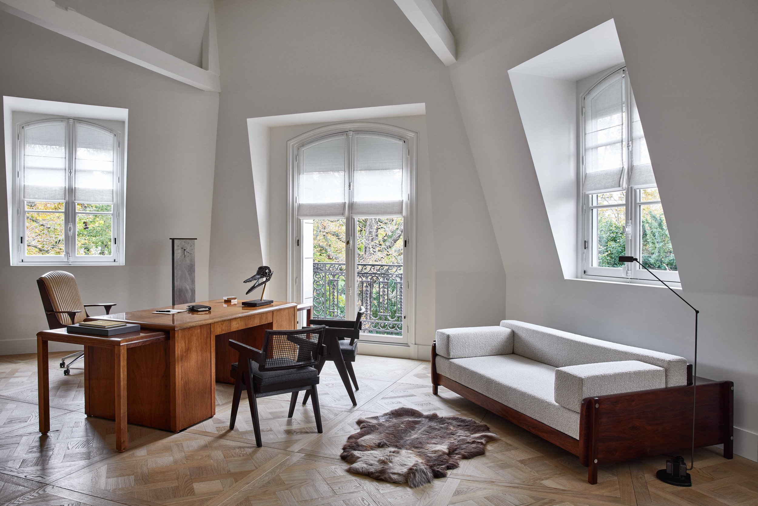 Un bureau moderne avec un grand bureau en bois, une chaise de bureau, un canapé blanc en bois de style scandinave, une étoffe de fourrure au sol, et des fenêtres lumineuses avec vue sur des arbres