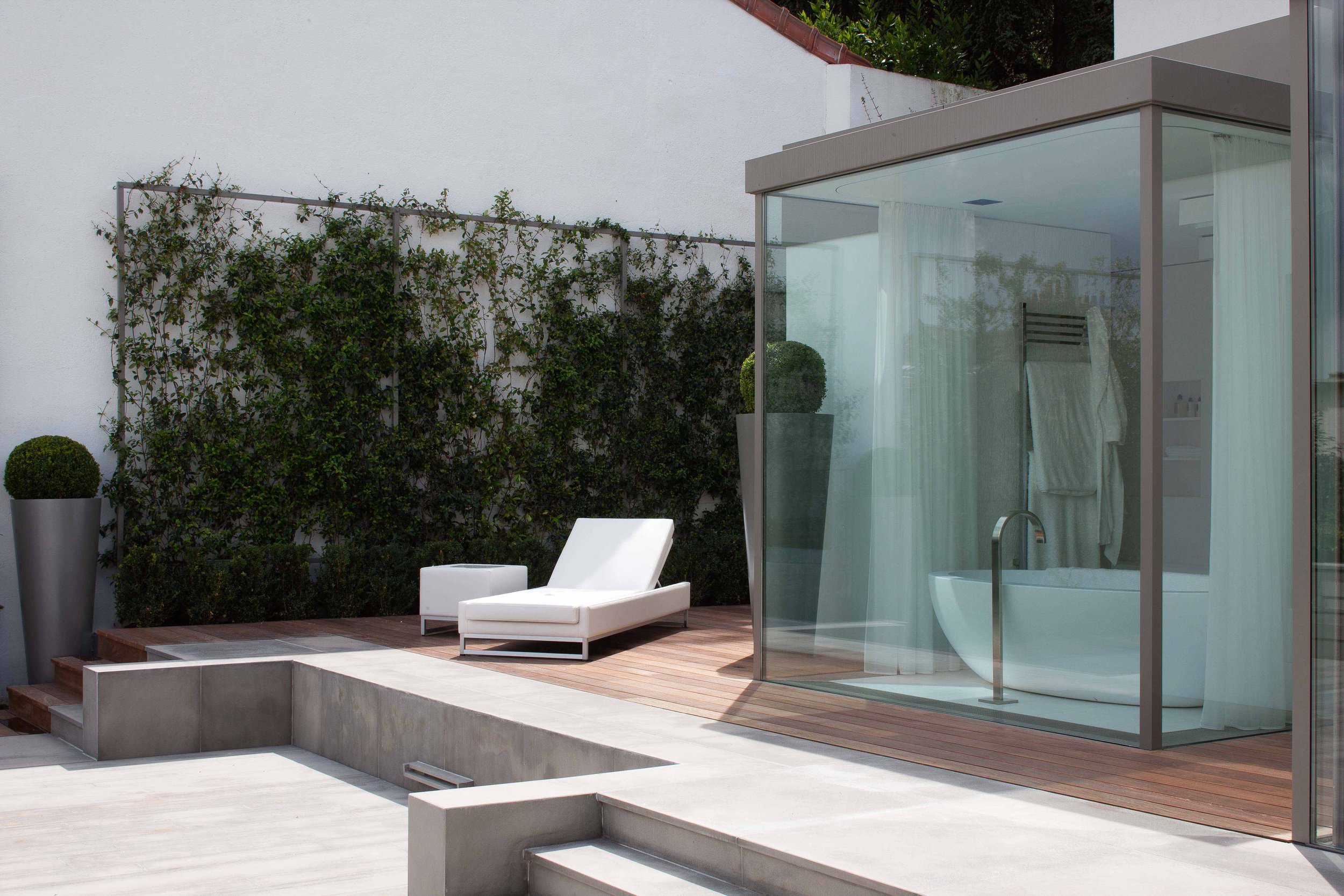 Une terrasse avec un lit de soleil blanc, une plante en pot, et une cabane en verre avec une baignoire et des serviettes à l'intérieur.