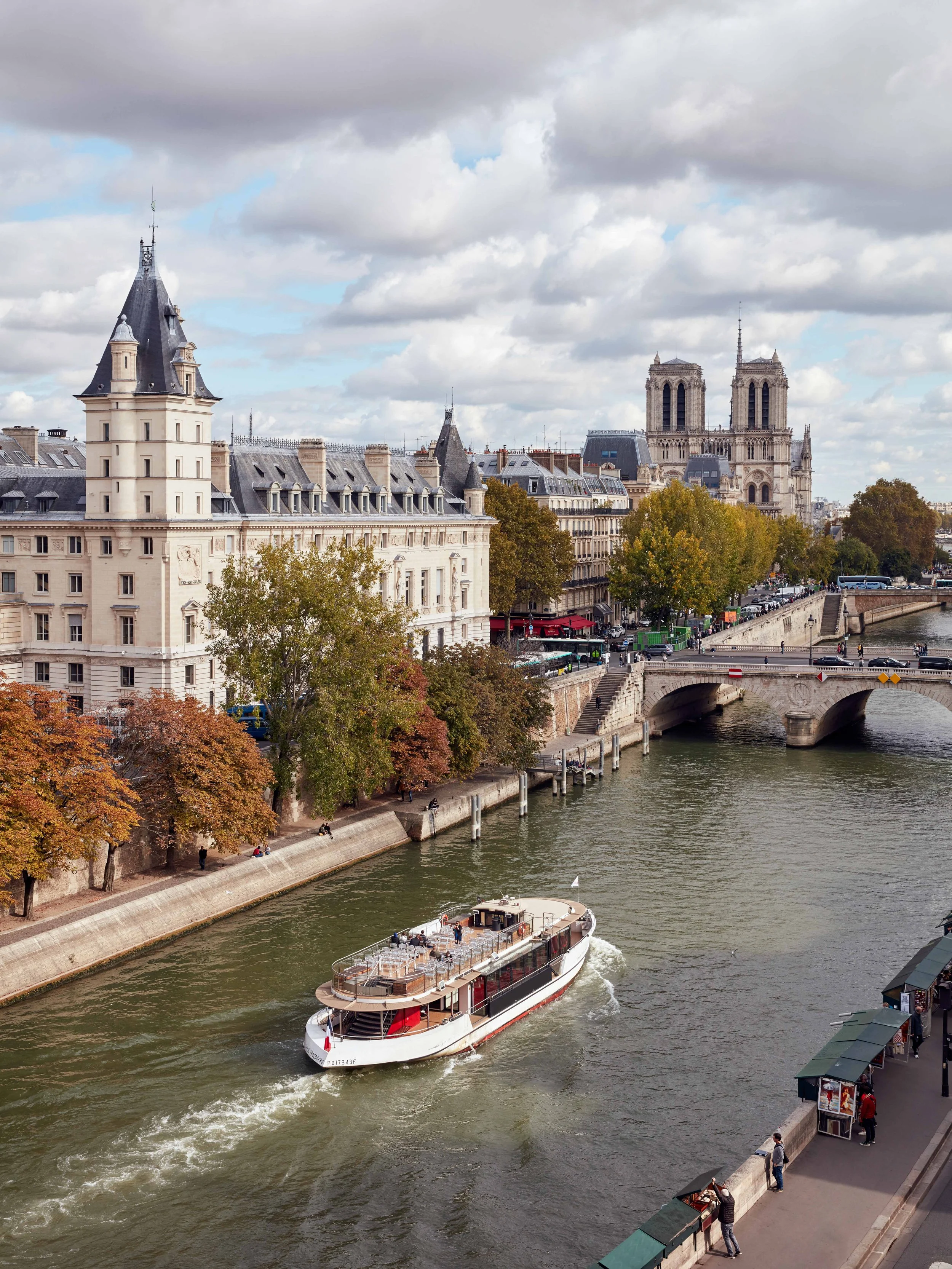 Vue d'une rivière à Paris avec un bateau touristique, des bâtiments historiques, des arbres aux couleurs d'automne, un pont piétonnier et la Cathédrale Notre-Dame en arrière-plan.