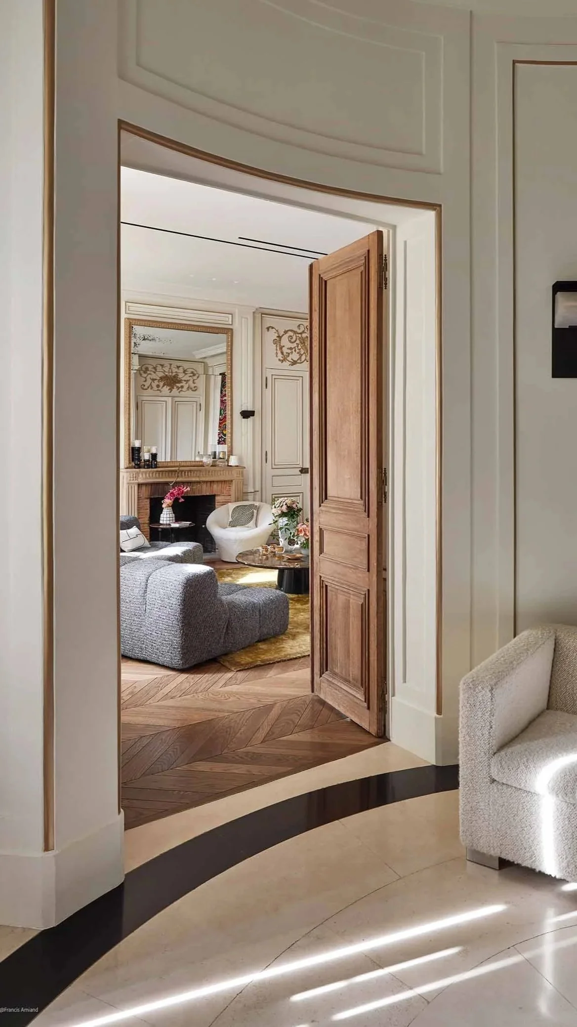 Une vue à travers une porte en bois ouverte dans un salon élégant, avec un canapé gris, une cheminée cornée, un miroir au mur, des fleurs sur une table basse et un fauteuil blanc dans une pièce riche en détails décoratifs.