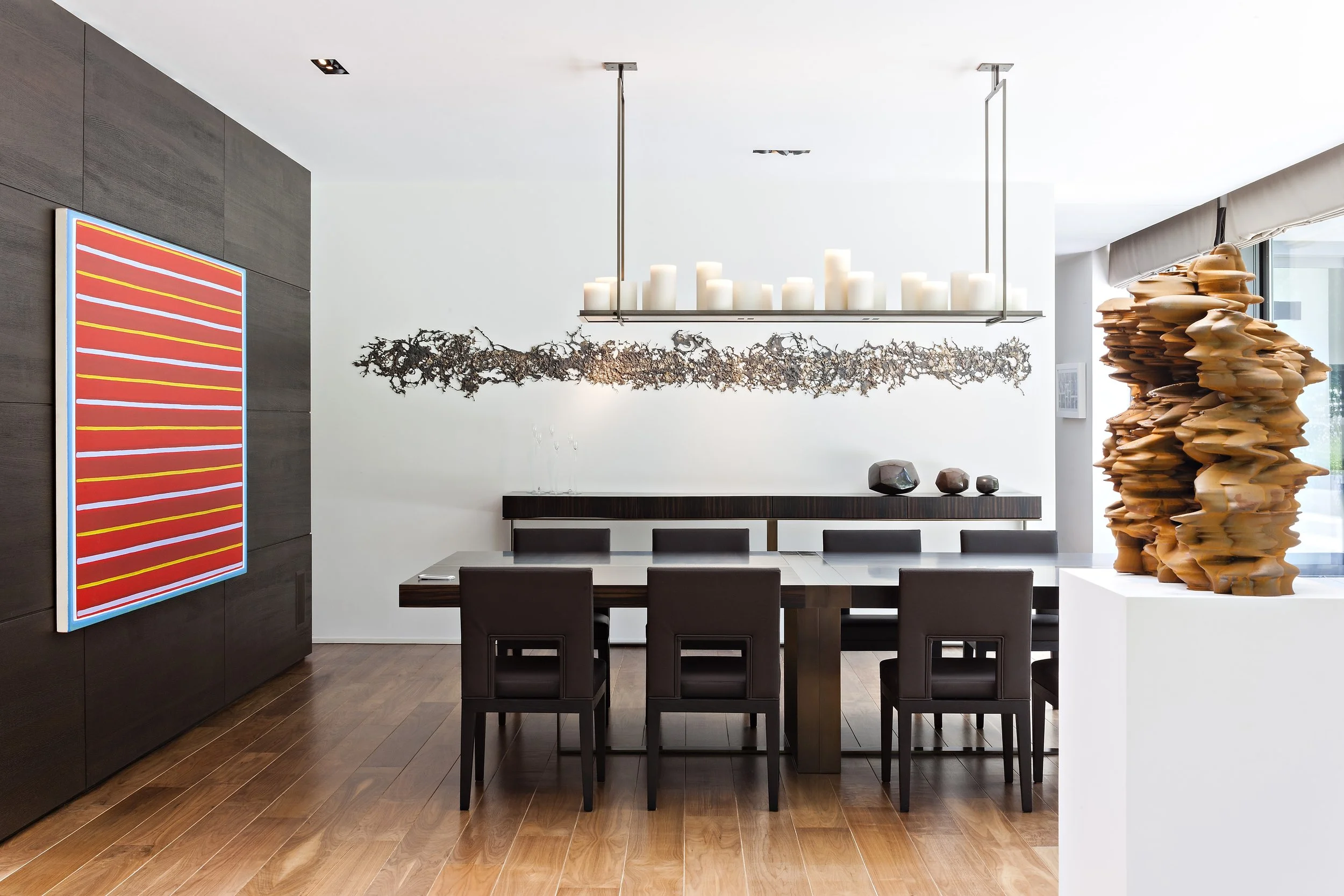 Salle à manger moderne avec une grande table, sept chaises en cuir noir, une œuvre d'art abstraite à gauche, une installation en bois sculpté à droite, et une suspension de bougies au plafond.