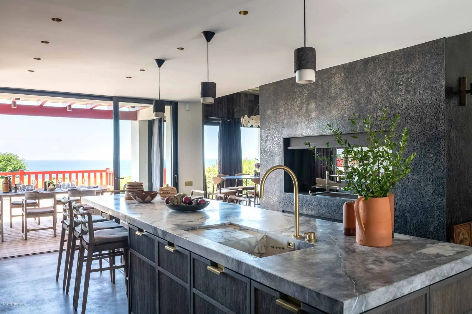 Cuisine ouverte avec un îlot central en marbre, un évier à gauche avec un robinet en or, une crasse de plantes dans un pot en terre cuite sur le comptoir, un mur en pierre noire et une grande fenêtre offrant une vue sur l'extérieur.