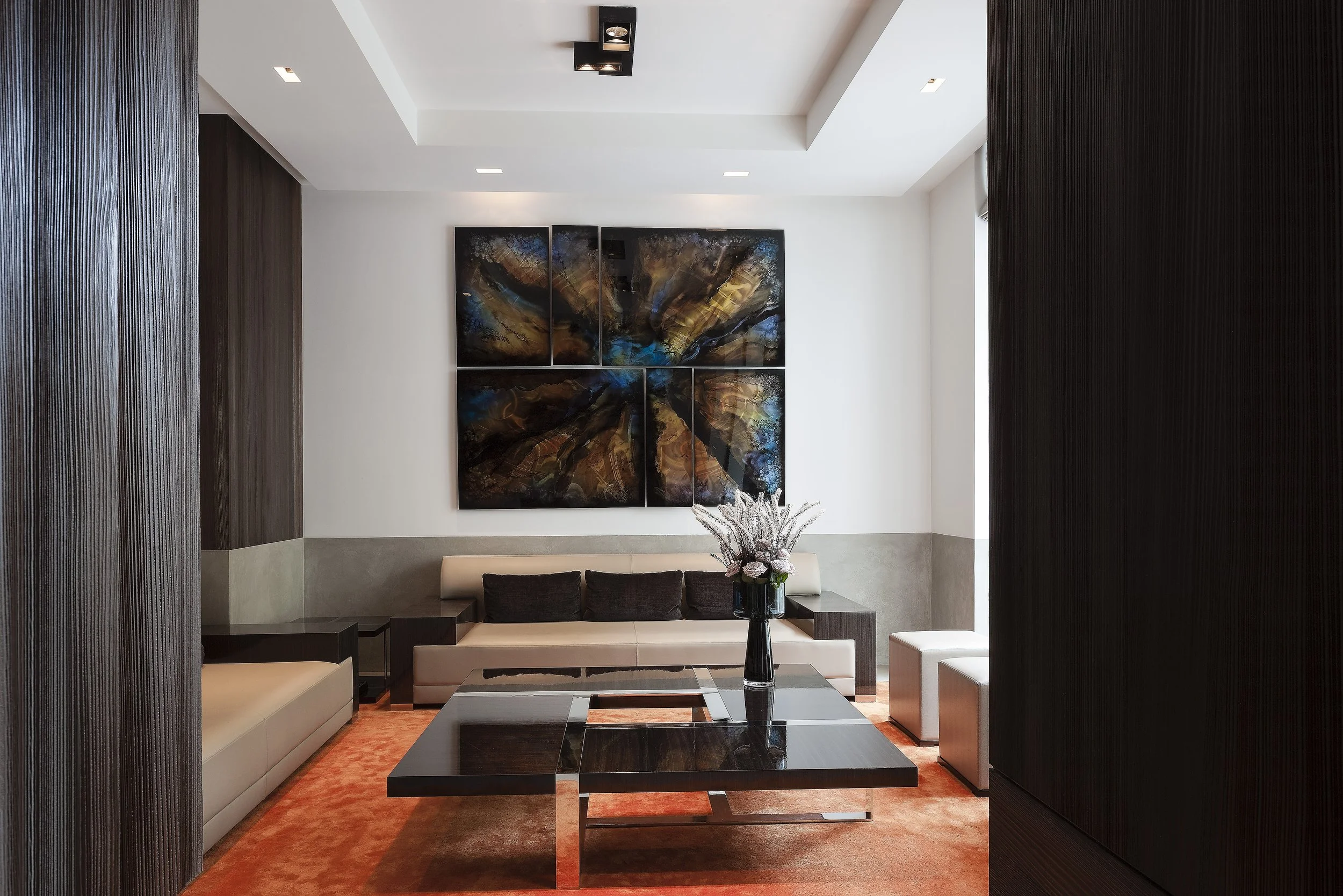 Salon moderne avec canapé beige, tableau abstrait coloré, table basse noire, vase avec fleurs, et tapis orange.