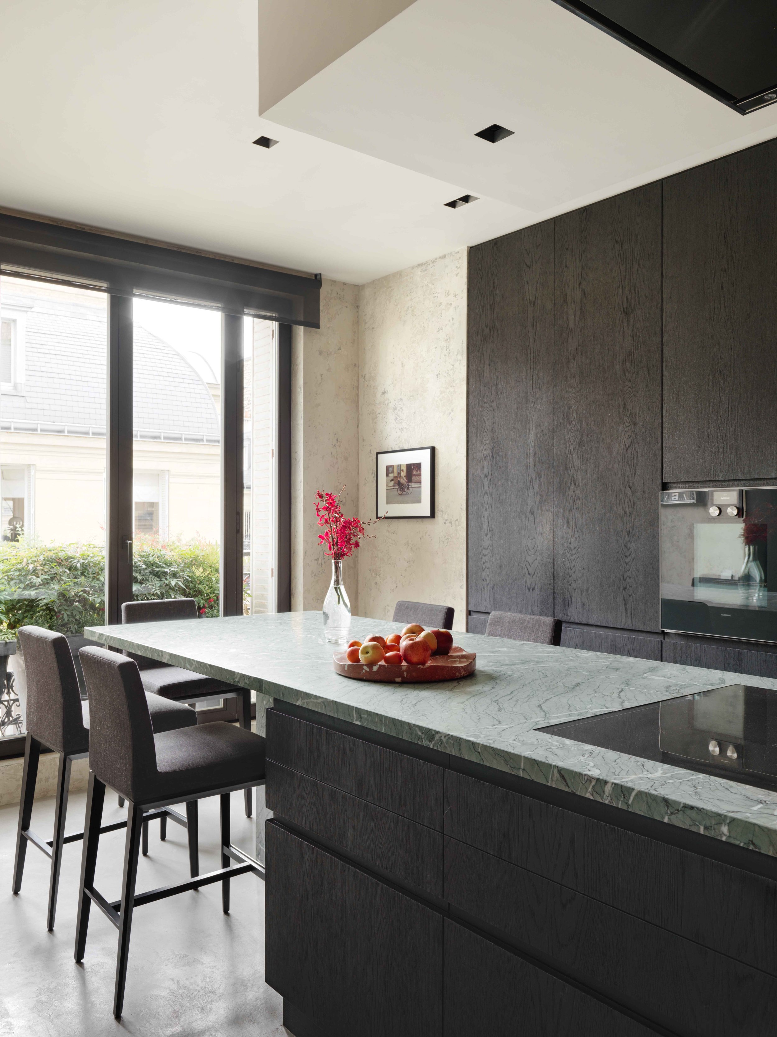 Cuisine moderne avec table en marbre vert, chaises en tissu gris, grande fenêtre, décoration minimaliste, tableau au mur, vase avec fleurs roses, fruits sur la table.