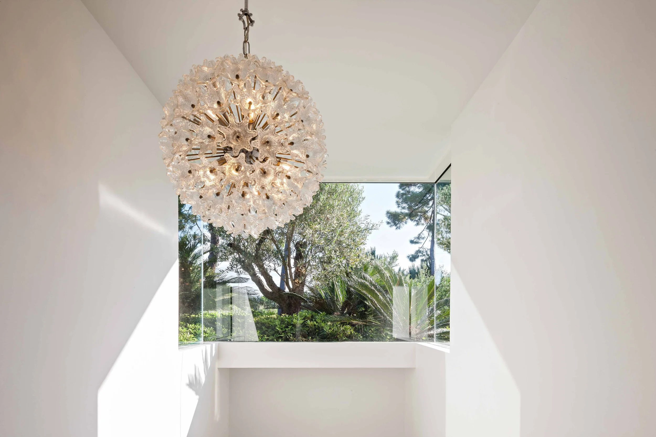 Un plafond avec un lustre en verre transparent dans une pièce blanche, avec une grande fenêtre donnant sur des arbres et du ciel