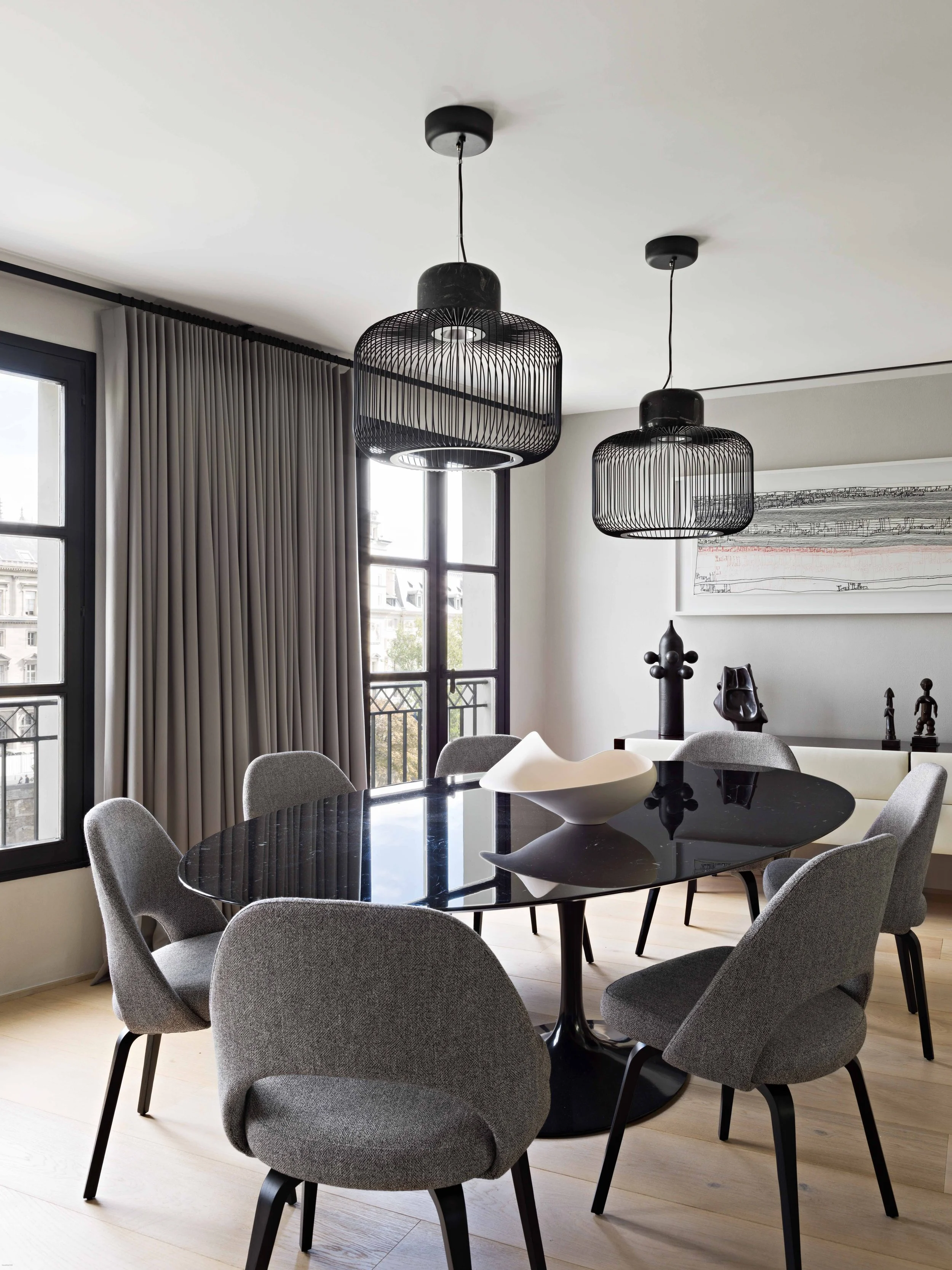 Salle à manger moderne avec table ronde en noir, six chaises gris, deux suspensions noires au plafond, grande fenêtre avec rideaux beiges, décoration artistique sur le mur.
