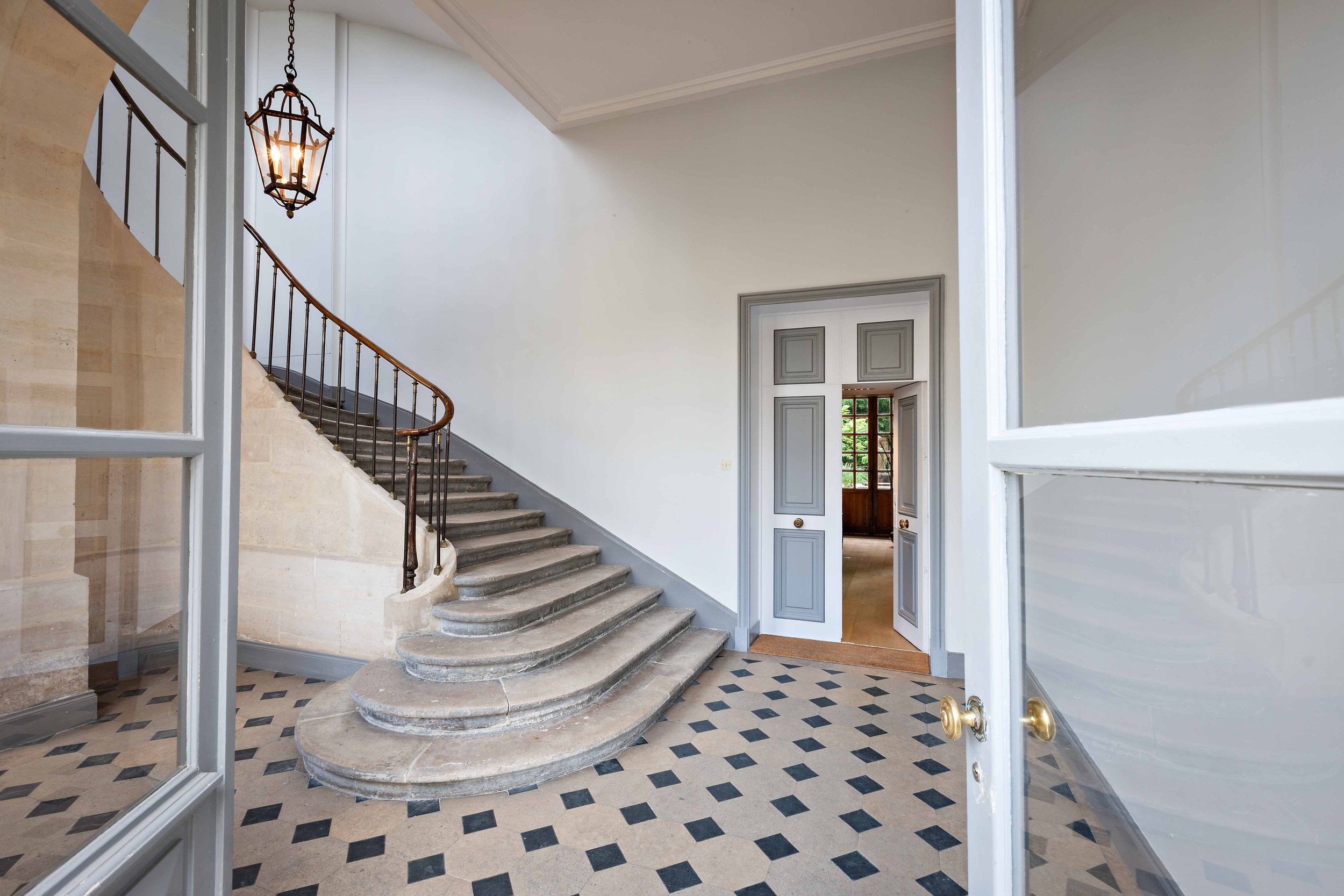 Intérieur d'une maison avec une escalier en pierre, garde-corps en fer forgé, portes en bois peint en gris, sol en carreaux de style classique, vue depuis une porte en verre avec poignée en laiton, espace lumineux avec un plafond blanc et une grille 