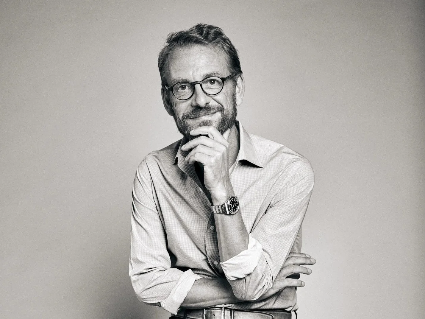 Homme âgé, avec lunettes, barbe et cheveux en bataille, pose en regardant pensivement, portant une chemise à manches retroussées, un bracelet en acier et une montre.