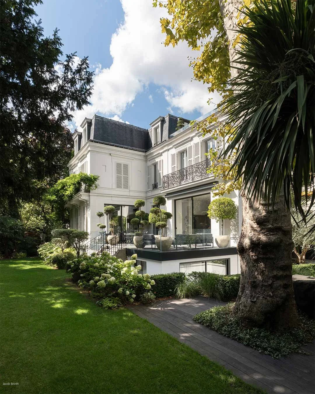 Maison blanche élégante avec jardin bien aménagé, plusieurs plantes en pot taillées en forme de boule, arbres et pelouse verte, sous un ciel bleu avec quelques nuages.