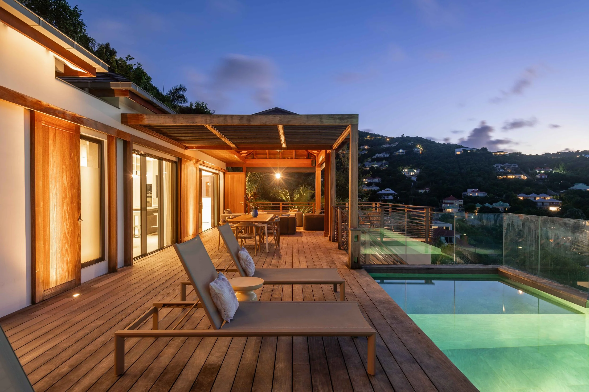 Terrasse en bois avec des chaises longues, une table, un jacuzzi, et un espace lounge, avec vue sur une colline à la tombée de la nuit.