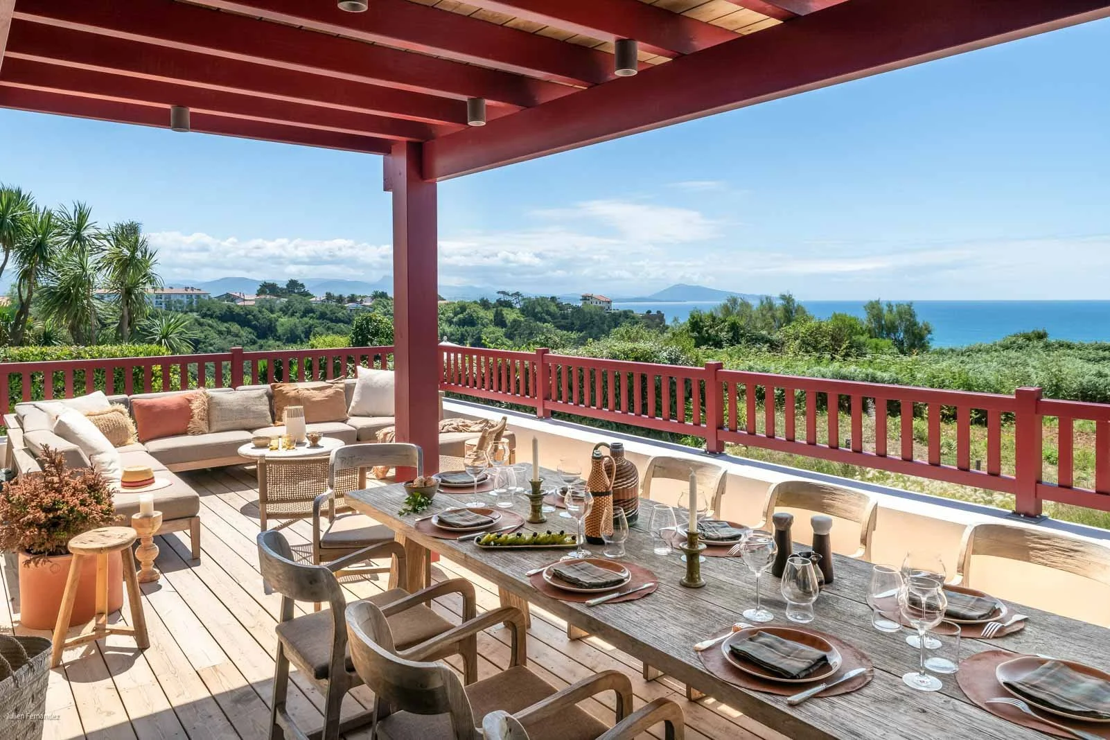 Terrasse avec mobilier de salle à manger, vue sur la mer, ciel ensoleillé, arbres et maison au loin.