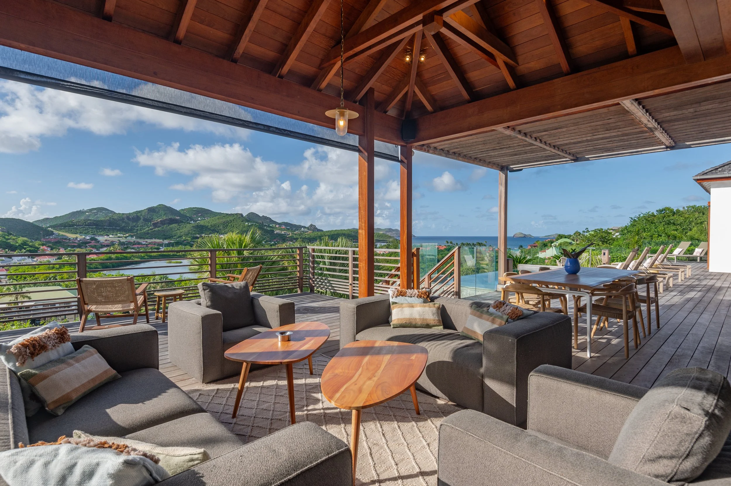 Terrasse avec vue panoramique sur la mer, montagnes et nature, mobilier de salon gris avec coussins, table basse en bois, terrasse en bois avec table à manger en bois et chaises, chaises longues en bois, ciel en partie nuageux.
