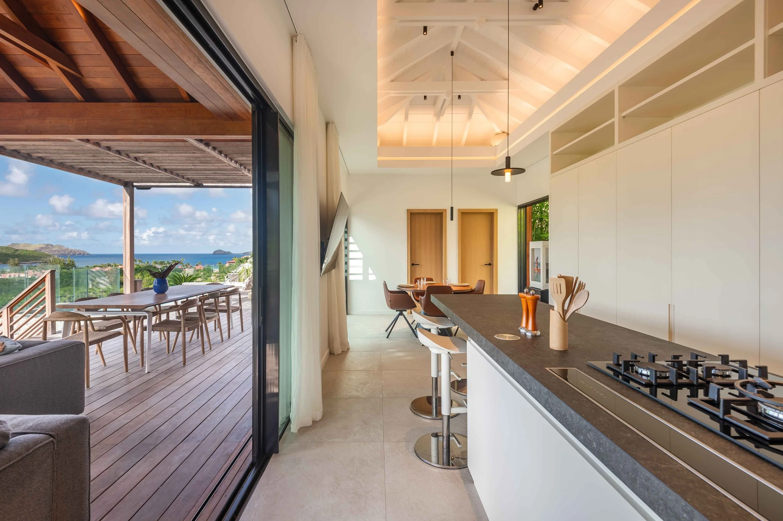 Intérieur d'une cuisine moderne en style minimaliste donnant sur une terrasse avec vue sur la mer, incluant un plan de travail en quartz noir, un évier, une table à manger en bois avec des chaises, et une porte en bois, avec un plafond en bois clair 