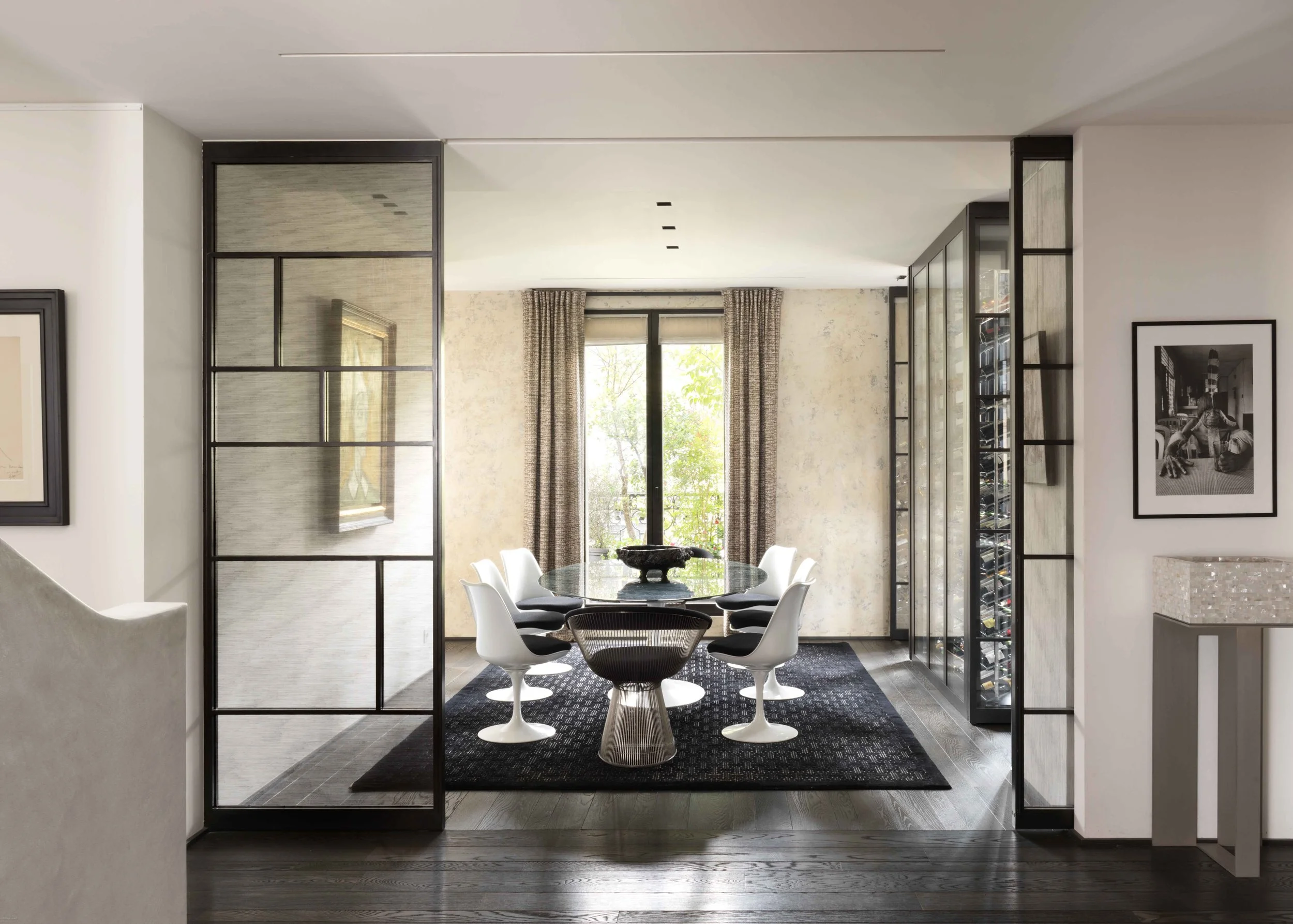 Salle à manger moderne avec une table en verre, huit chaises noires et blanches, et un grand mur de fenêtres laissant entrer la lumière naturelle. Décor épuré avec tapis noir et murs clairs.