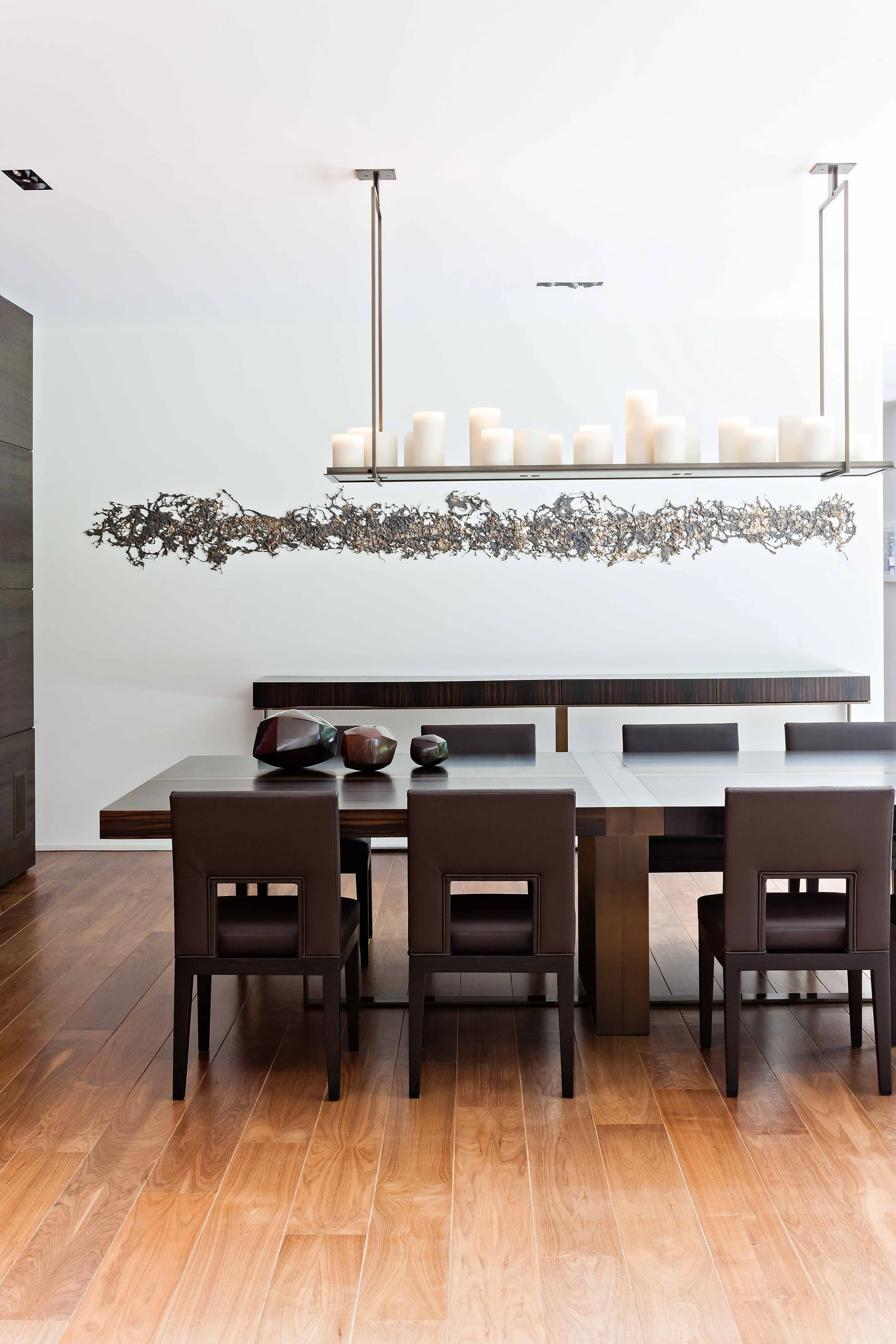 Salle à manger moderne avec une grande table en bois foncé, six chaises en cuir marron, un chandelier suspendu avec des bougies, et une décoration murale artisanale.