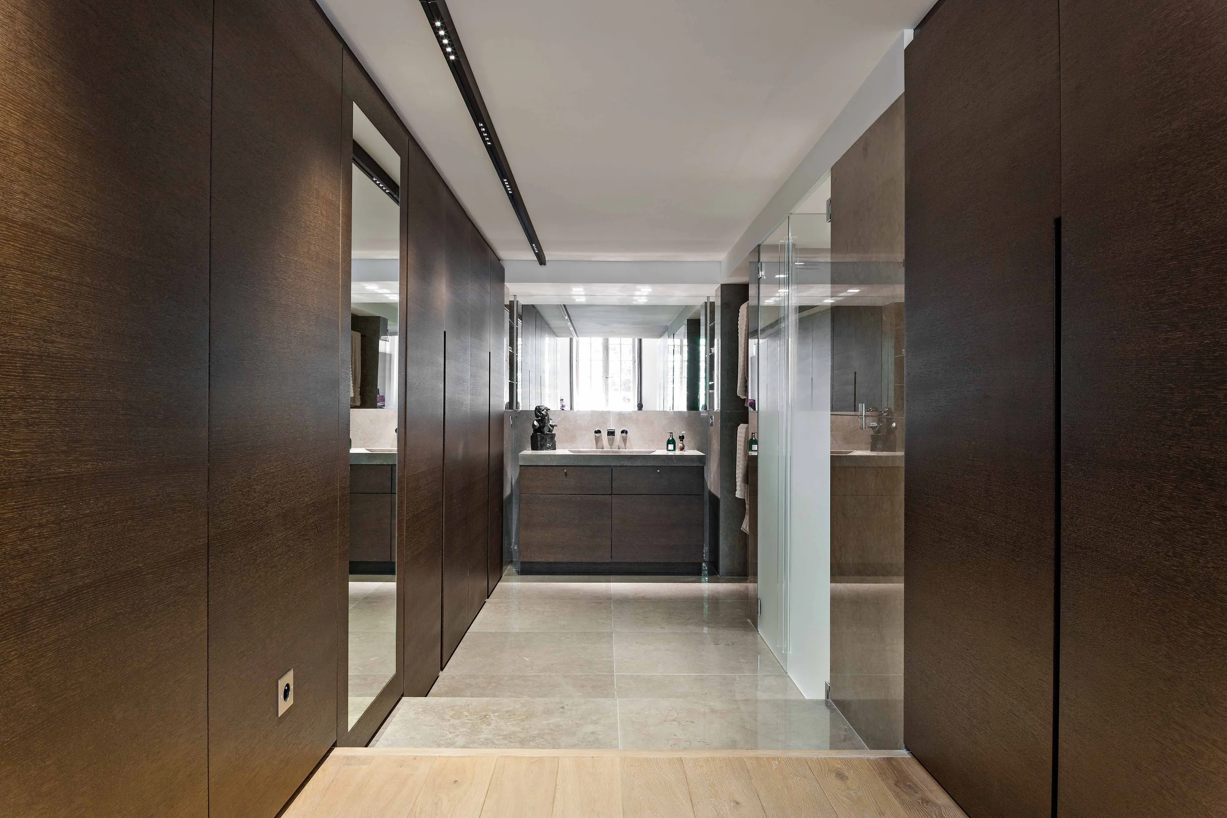 Intérieur d'une salle de bain moderne avec murs en bois foncé, sols en pierre claire, grande fenêtre laissant entrer la lumière naturelle, lavabo en pierre avec miroir au plafond, quelques objets de décoration sur le lavabo, paroi en verre séparant l