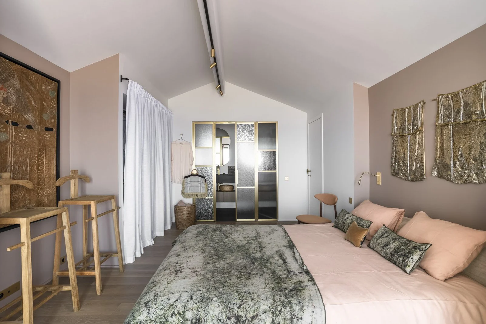 Chambre décorée avec un grand lit à draps roses, coussins en velours gris et or, mur rose pâle, porte en bois clair, miroir, et séparation en verre opaque avec cadres dorés.