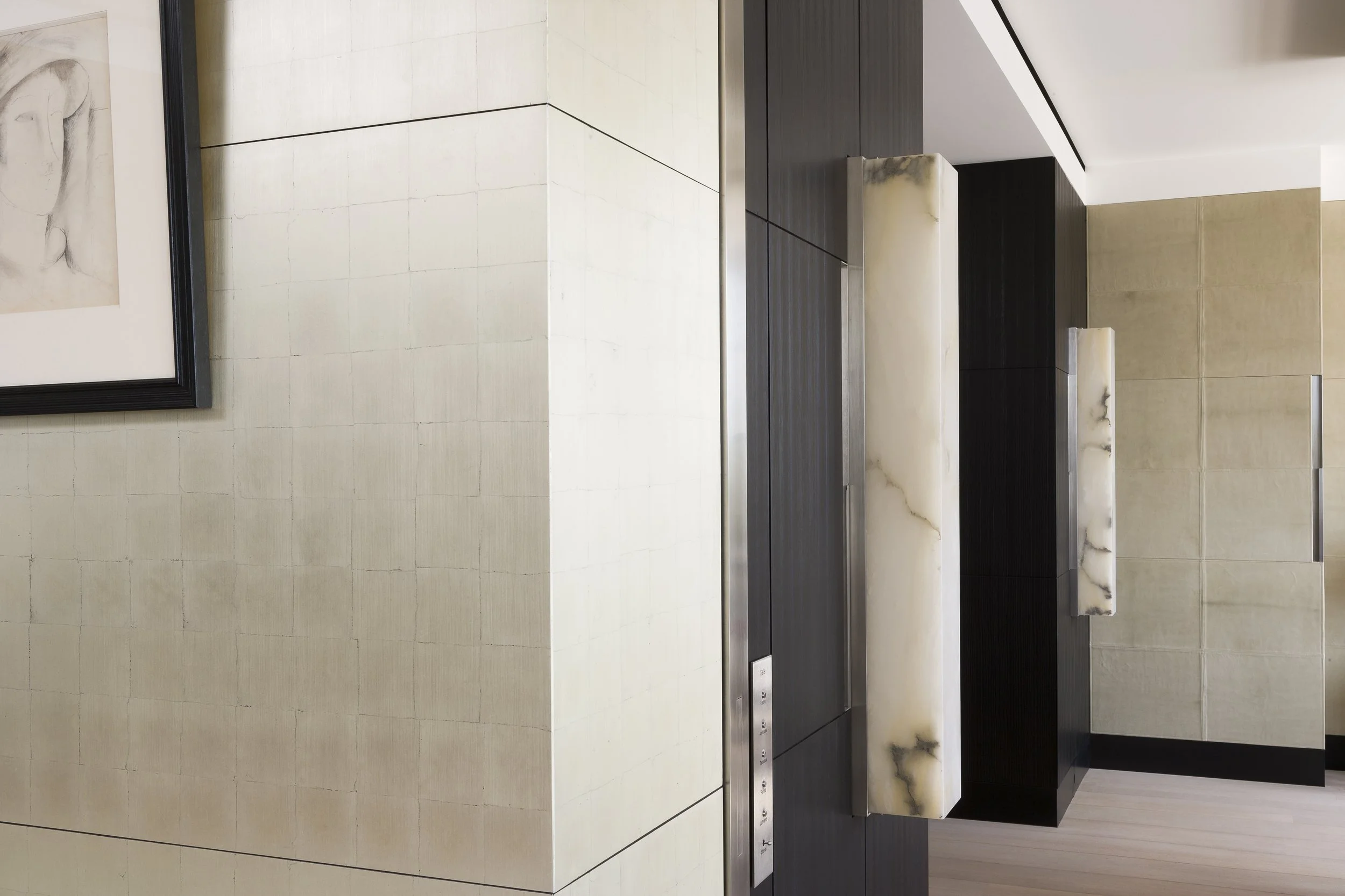 Intérieur d'un bâtiment moderne avec murs en pierre beige clair, une œuvre d'art encadrée et un ascenseur avec boutons visibles