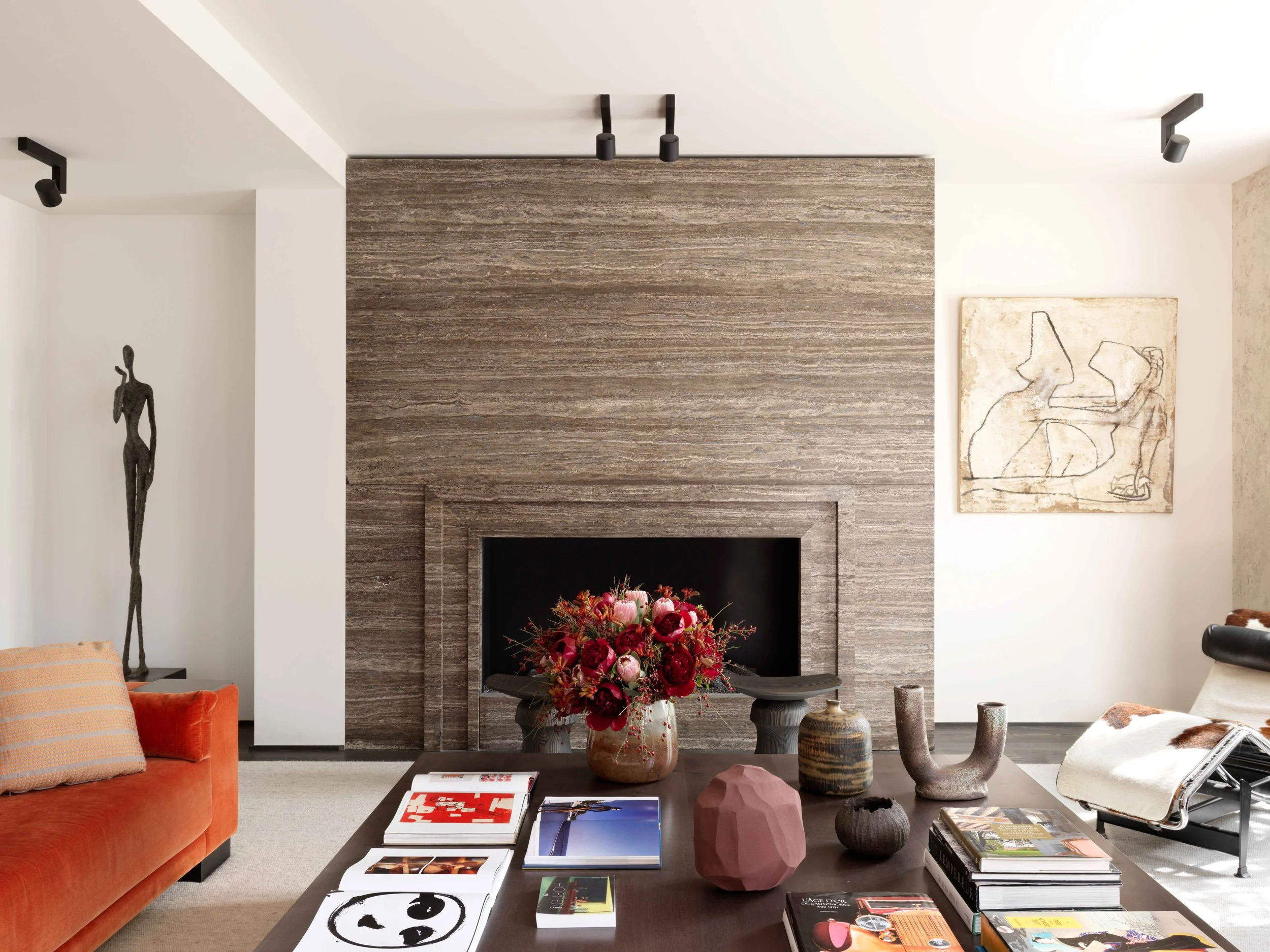 Un salon moderne avec une cheminée en pierre, un canapé orange, une table basse avec des livres, des vases et un bouquet de fleurs, et des œuvres d'art murales.