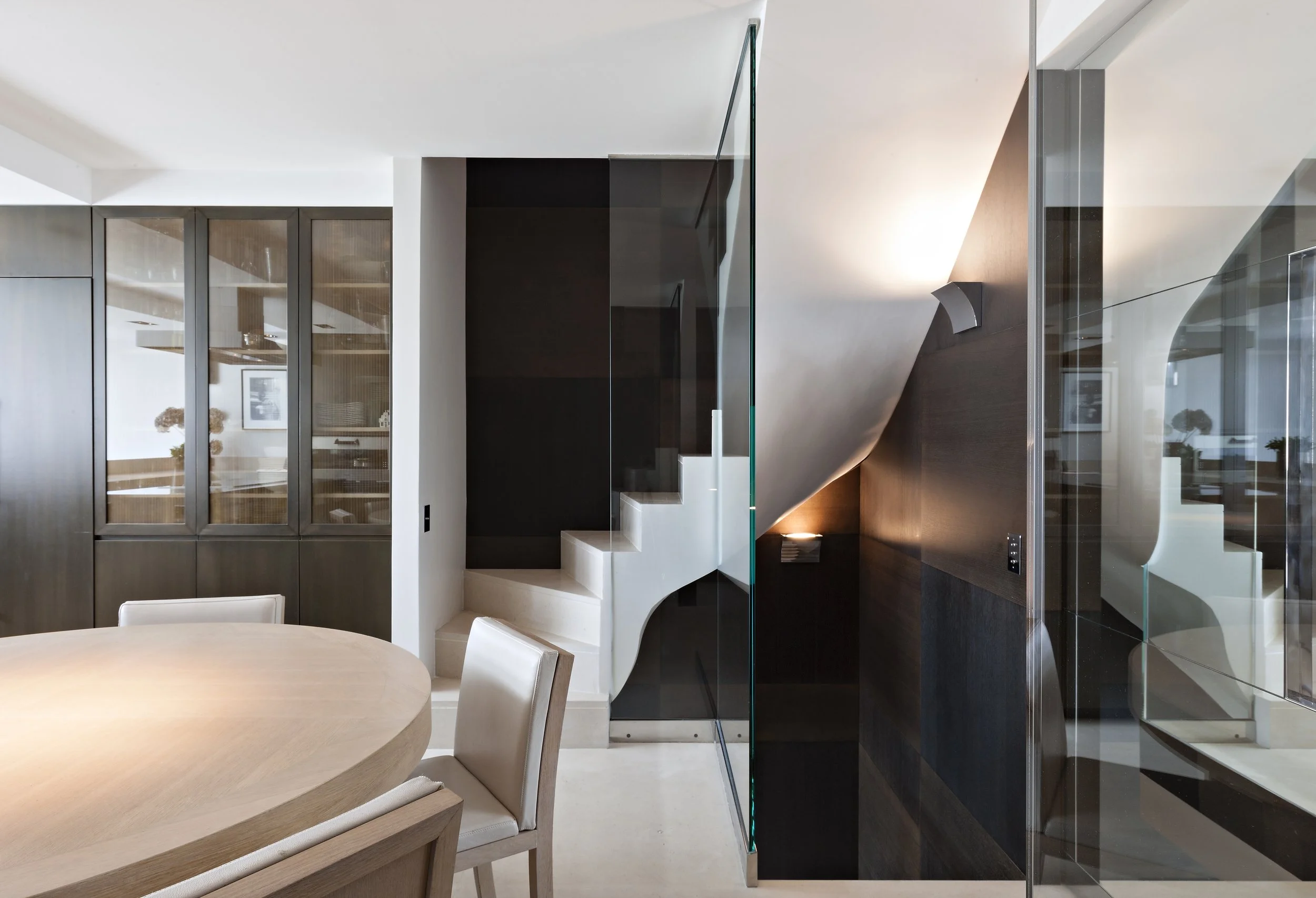 Intérieur moderne d'une salle à manger avec une table ronde en bois, des chaises en tissu beige, un mur en miroir, un escalier blanc avec une paroi en verre, et un plafond blanc avec éclairage.