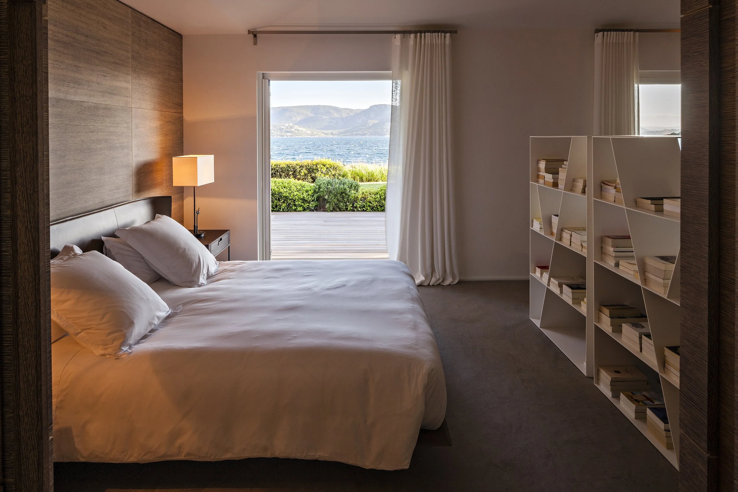 Chambre moderne avec lit king-size, vue sur l'eau, étagère blanche remplie de livres, porte-fenêtre avec rideau blanc donnant sur un terrasse extérieure