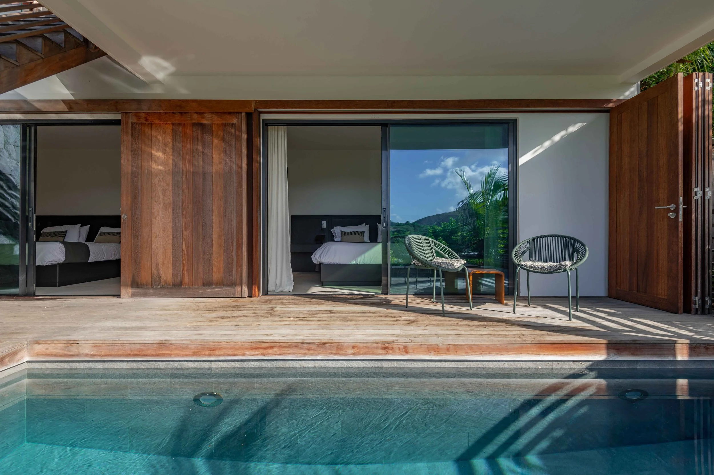 Terrasse d'une maison avec piscine, deux chaises en rotin avec coussins, vue sur une chambre avec lit et vue sur la nature, intérieur moderne avec murs en bois et grandes fenêtres en verre.