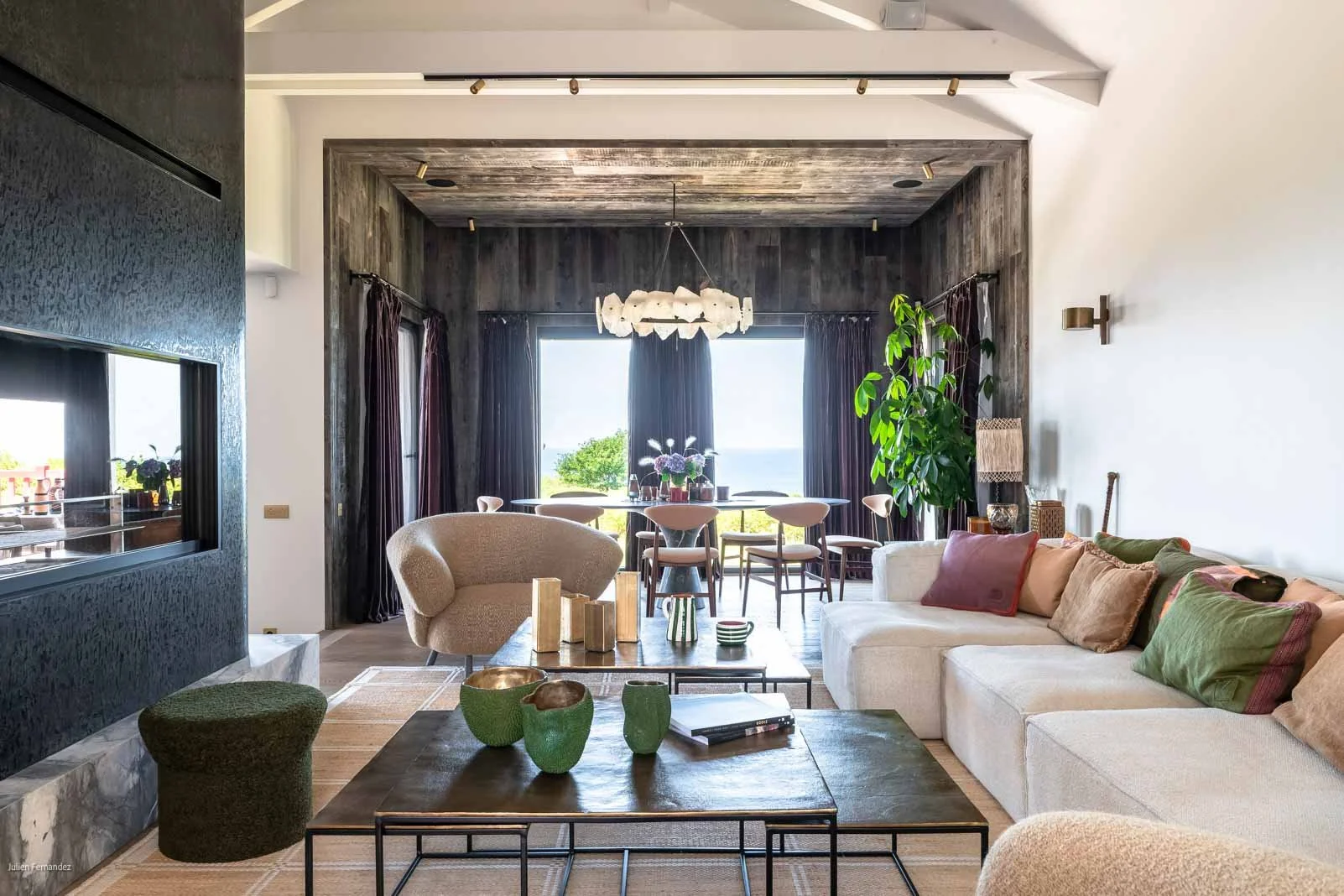 Salon moderne avec canapé blanc, coussins colorés, tables en bois, et vue sur l'extérieur par une grande fenêtre