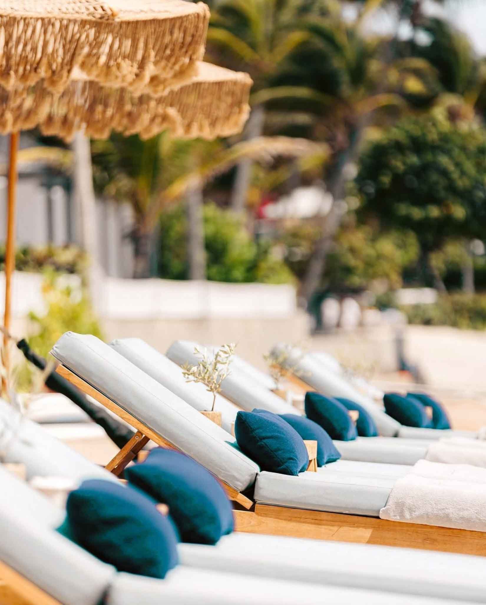 Chaises longues avec coussins bleus sous des parasols en paille, en extérieur, avec des arbres verdoyants en arrière-plan.
