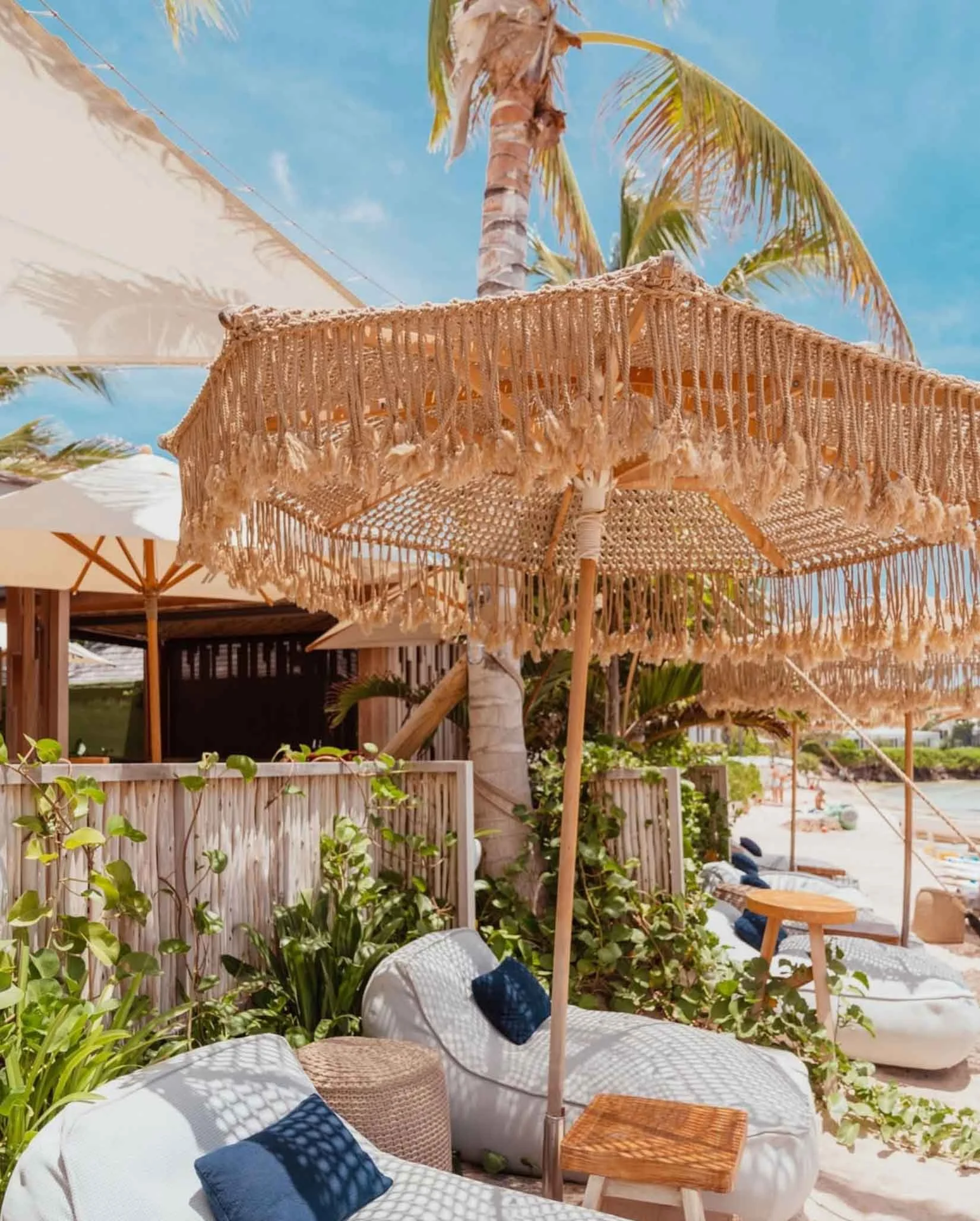 Un parasol en rotin tressé avec des franges, placé sur une plage ensoleillée, entouré de chaises longues avec des coussins et de palmiers en toile de fond.