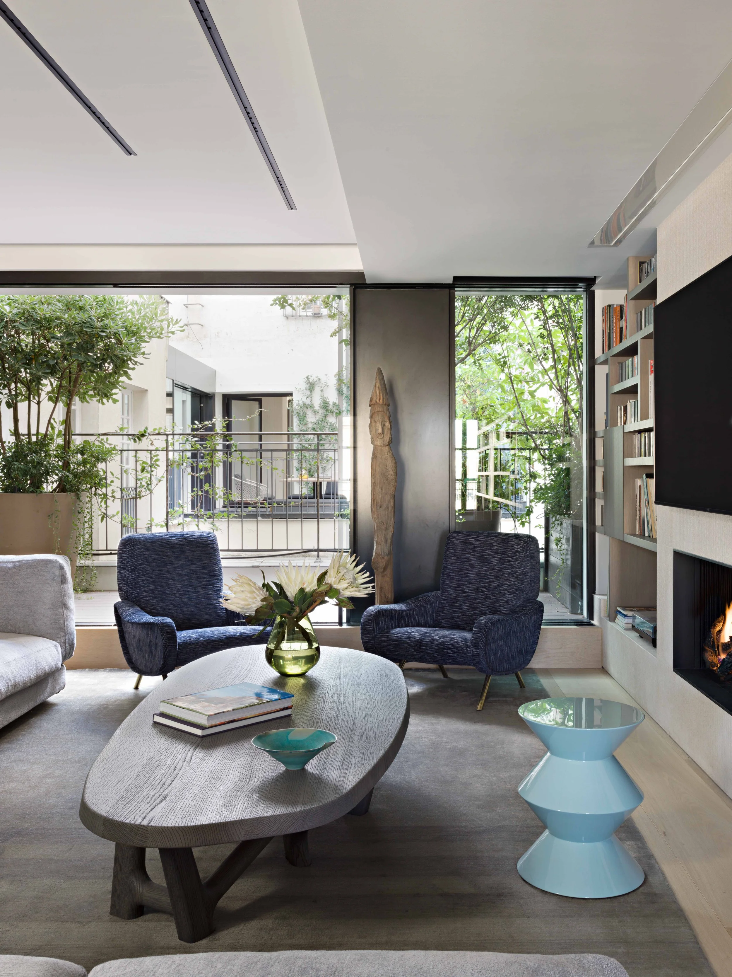 Intérieur d'un salon moderne avec deux fauteuils bleus, un canapé gris, une table basse en bois avec un vase de fleurs, un livre et un bol, une cheminée allumée, étagère avec des livres, grand mur en verre avec vue sur un balcon avec des plantes et d