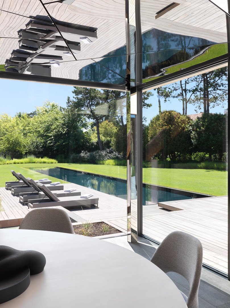 Vue intérieure d'une salle donnant sur un jardin avec piscine, chaises longues et arbres, vue à travers de grandes fenêtres en verre.