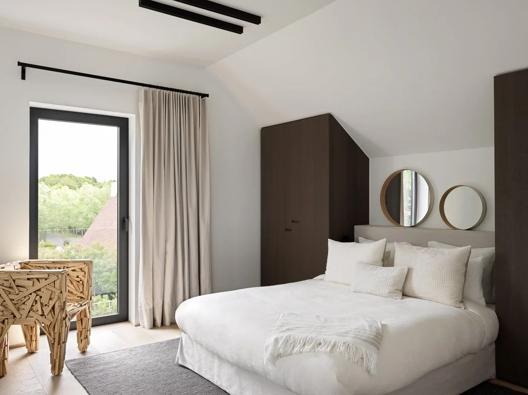 Chambre moderne avec lit blanc, deux miroirs ronds au-dessus, armoire en bois, grande fenêtre avec rideaux, siège en bois à motifs, tapis gris.