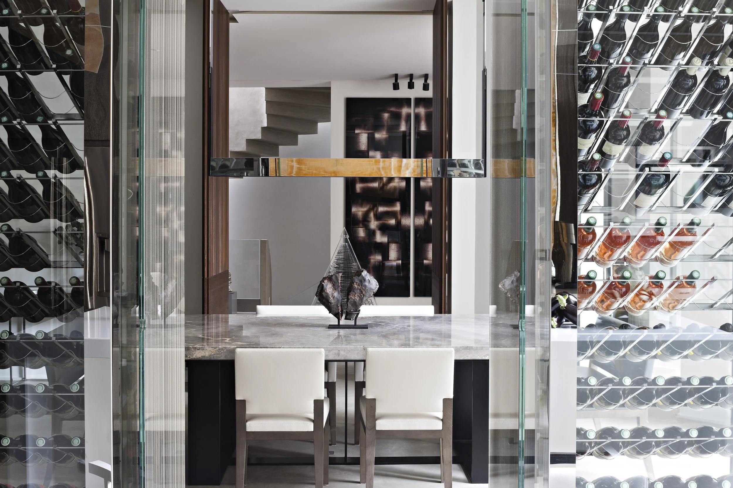 Vue intérieure d'une cave à vin moderne avec rayonnages de bouteilles en verre, un mur noir abstrait, une table en marbre avec deux chaises blanches, et une sculpture en métal sur la table.