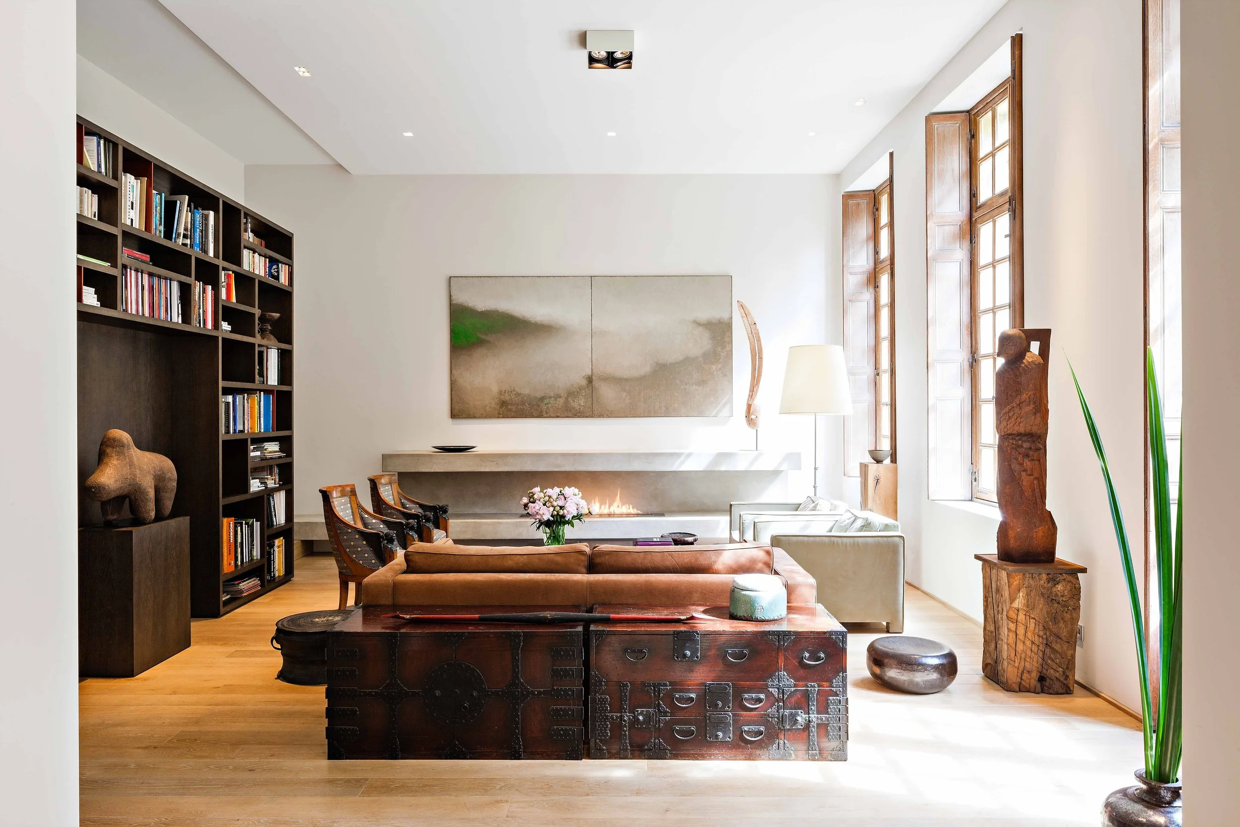 Salon moderne avec cheminée, étagère remplie de livres, sculptures en bois, fauteuils, canapé, et décoration intérieure lumineuse.