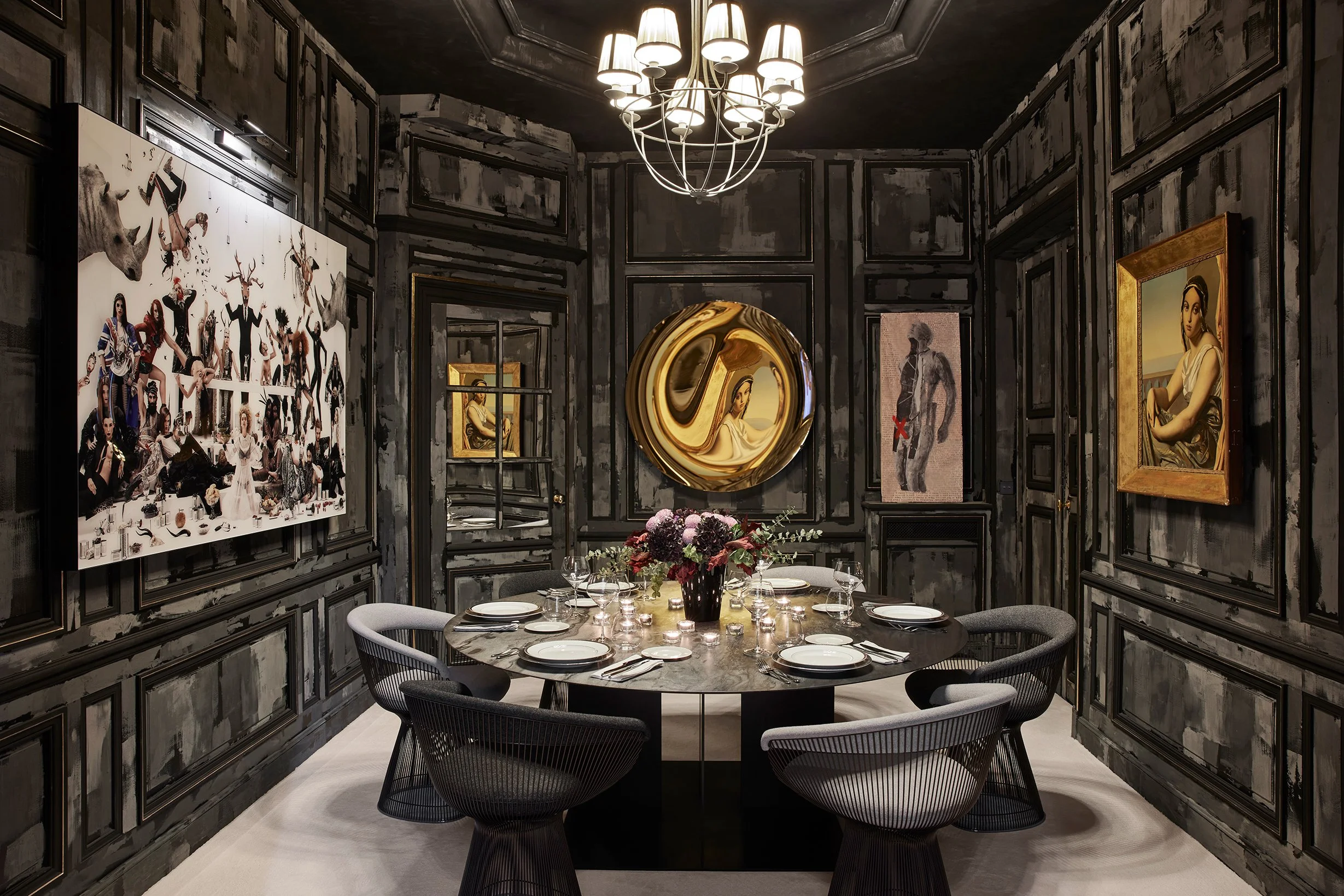 Salle à manger élégante avec murs sombres, un grand vase de fleurs au centre, une table ronde dressée avec des assiettes, verres et couverts, et des œuvres d'art modernes accrochées aux murs.