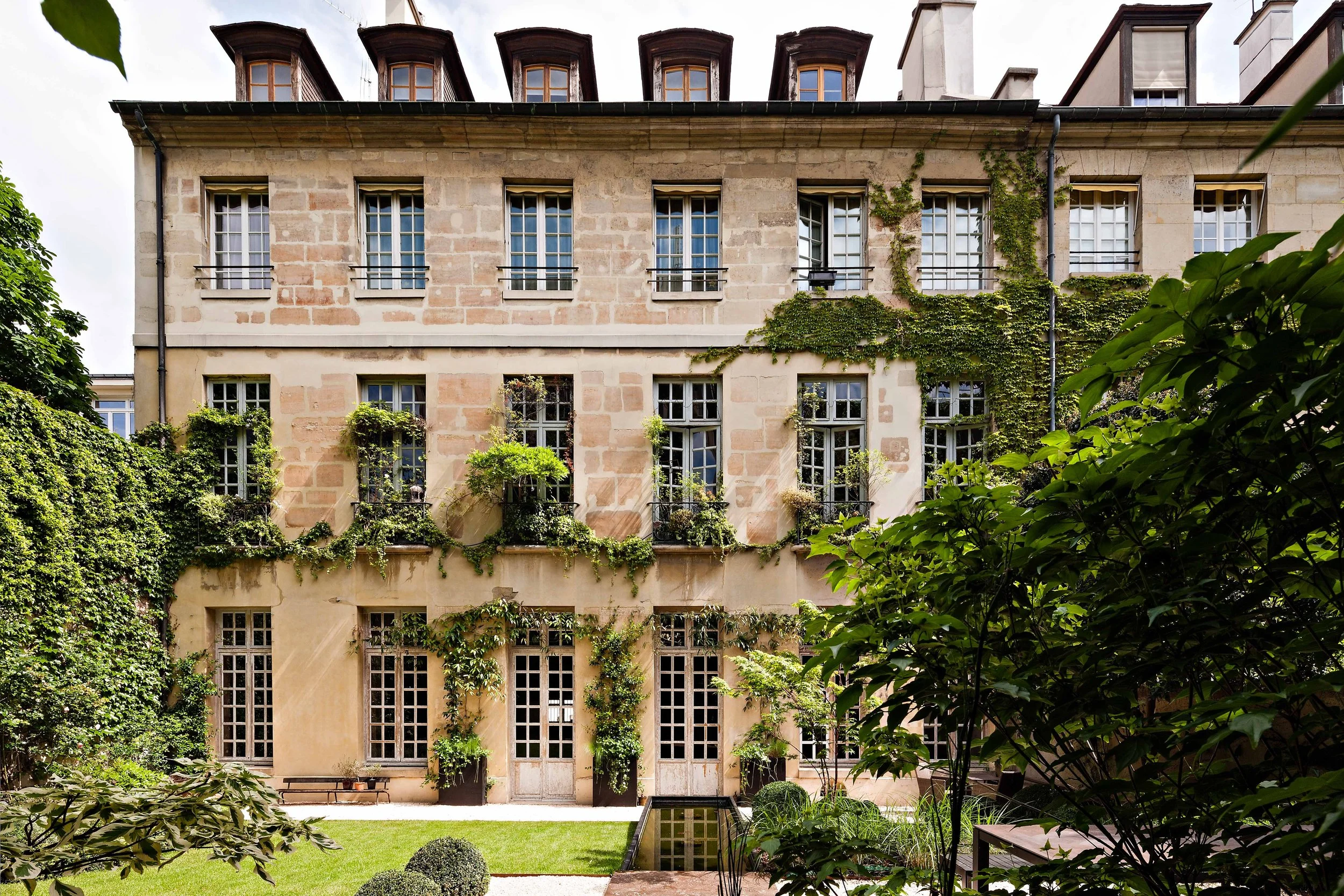 Vue d'une façade d'immeuble en pierre avec plusieurs fenêtres, des balcons avec des plantes vertes, et un jardin bien entretenu avec des buissons et de l'herbe.