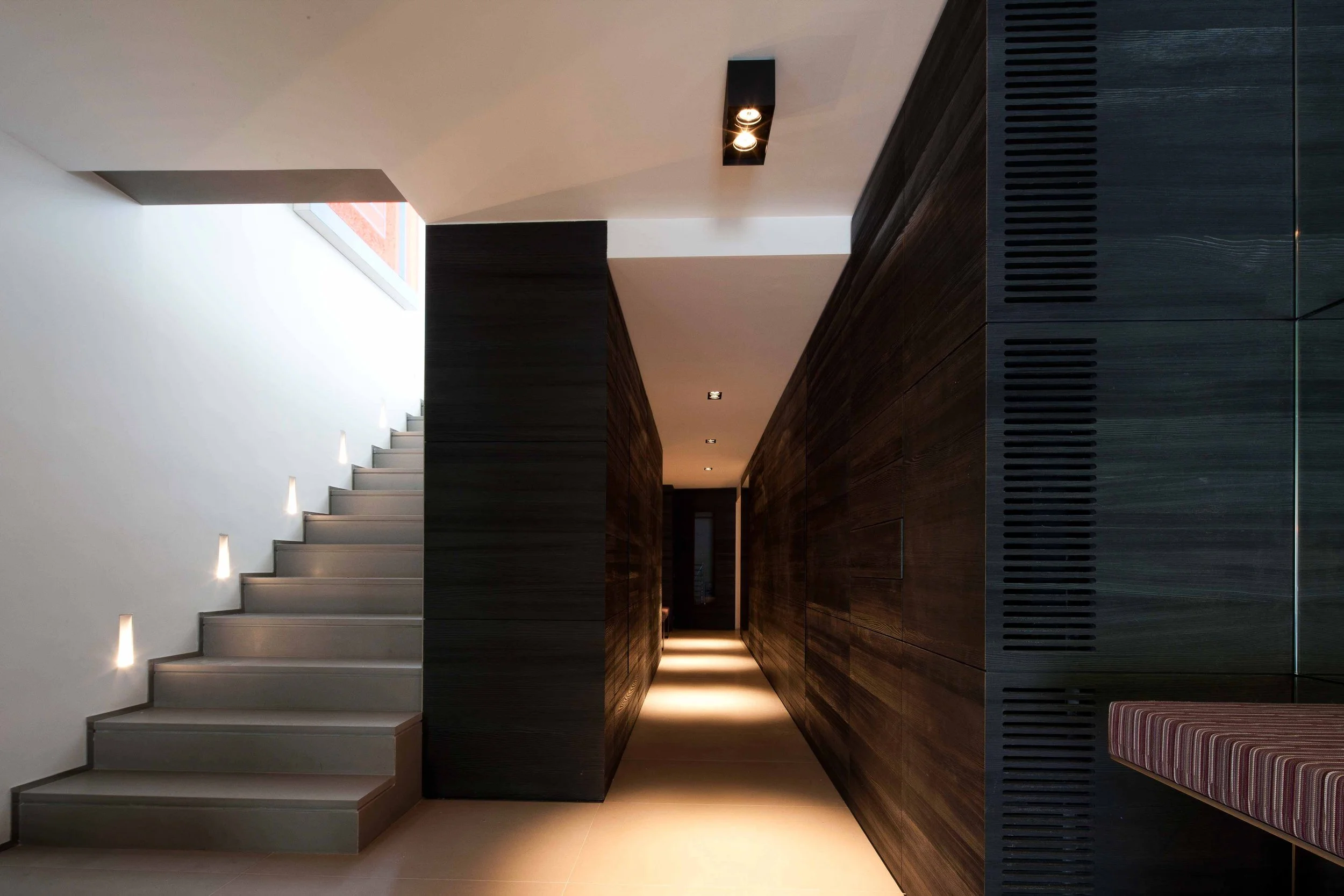 Couloir intérieur moderne avec escalier en acier, murs en bois foncé et éclairage encastré au sol.