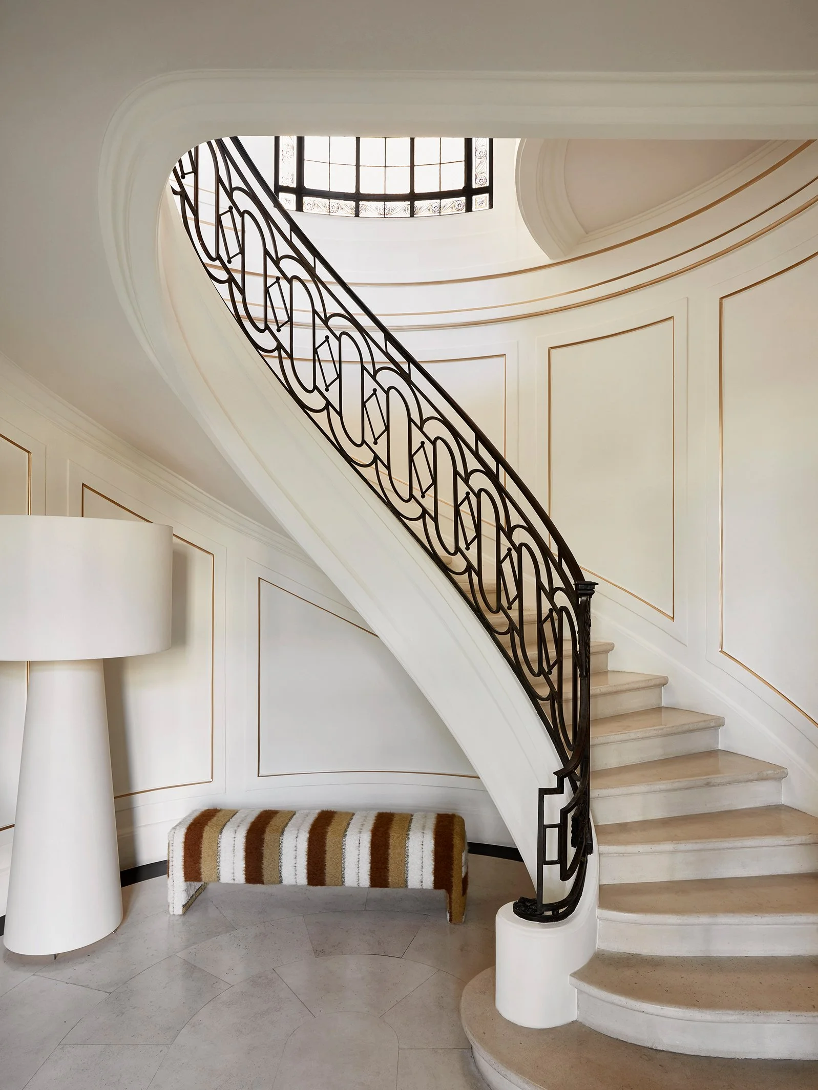 Escalier en colimaçon avec rampe en fer forgé, mur blanc avec moulures, lampe de sol blanche et banc rembourré à rayures marron, beige et blanc.