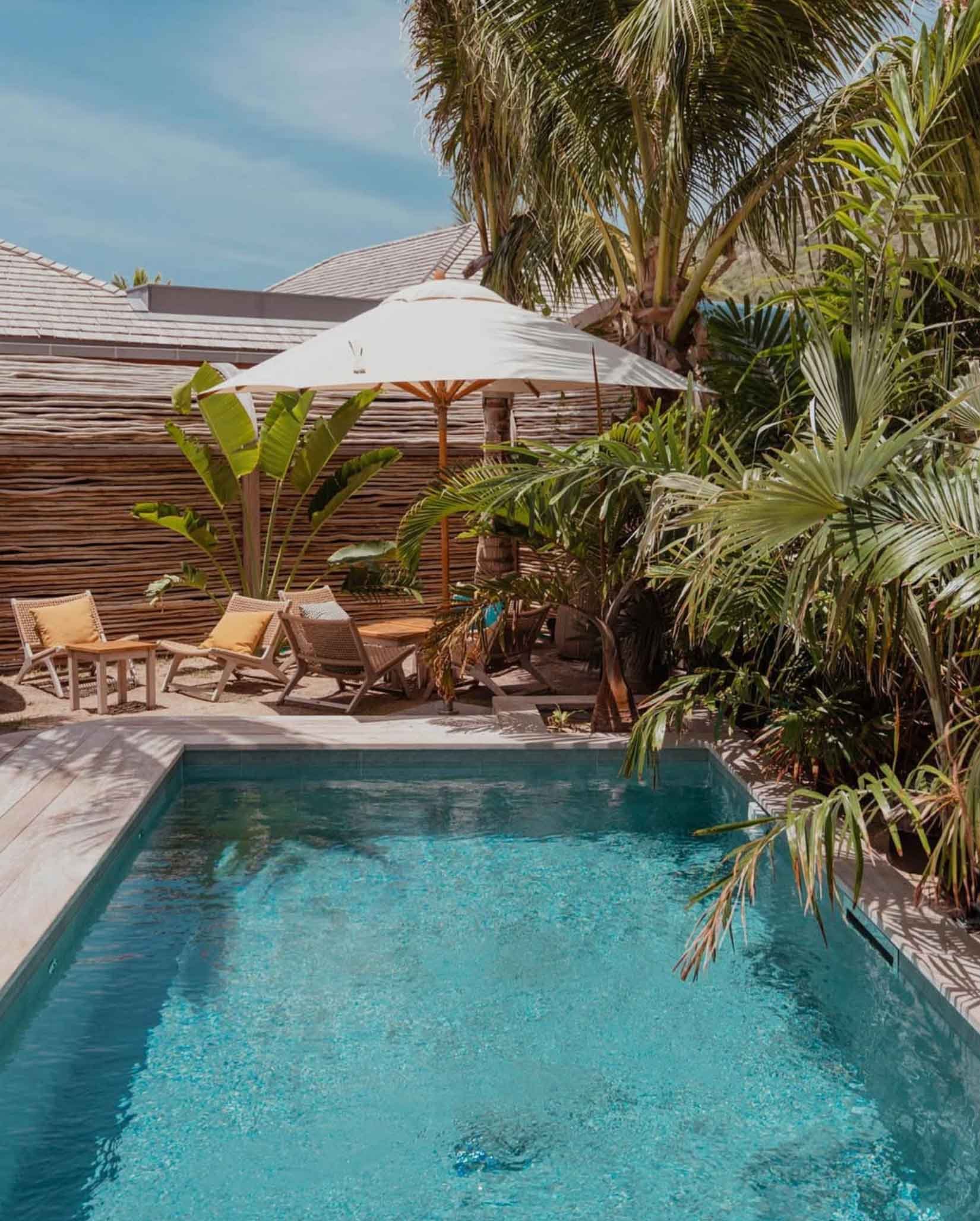 Piscine extérieure entourée de chaises longues avec coussins, parasol blanc, et plantes tropicales, avec un mur en bois et un toit en tuiles en arrière-plan.