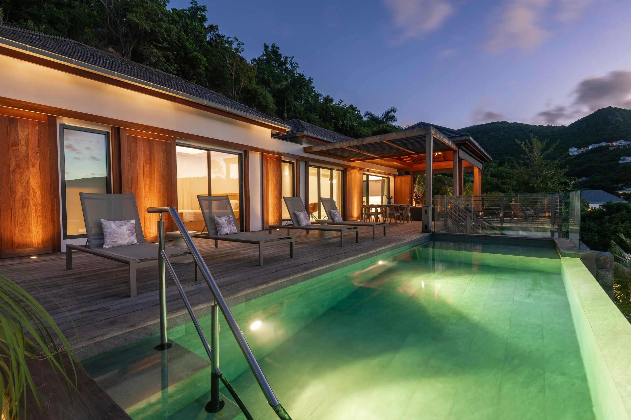 Une maison moderne avec une piscine éclairée, terrasse en bois avec des fauteuils et une table. La maison est située dans un environnement montagneux avec des arbres et des maisons en arrière-plan au crépuscule.
