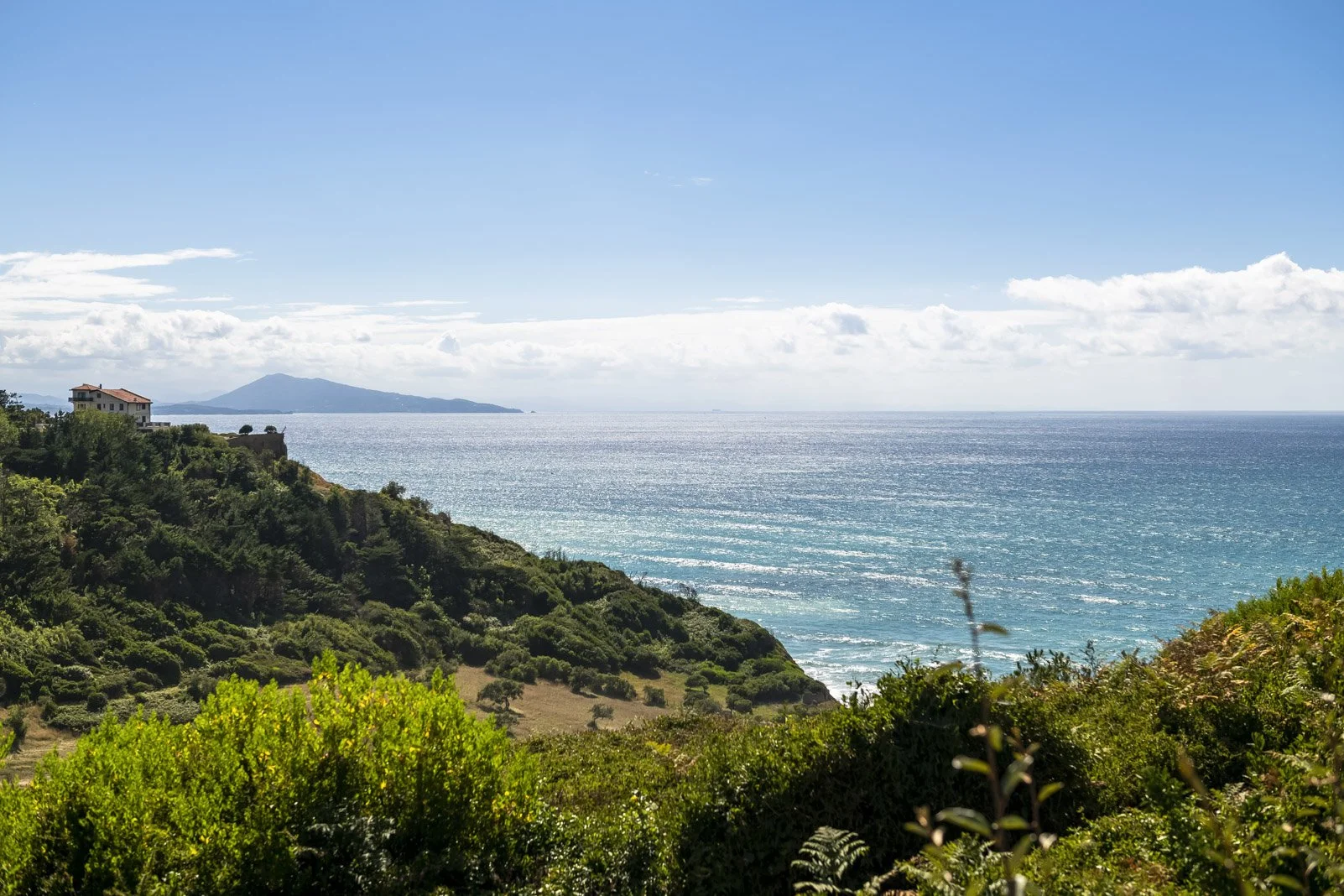 Paysage côtier avec mer bleue, collines verdoyantes, maison sur une falaise, île ou montagne au loin, ciel bleu avec quelques nuages.
