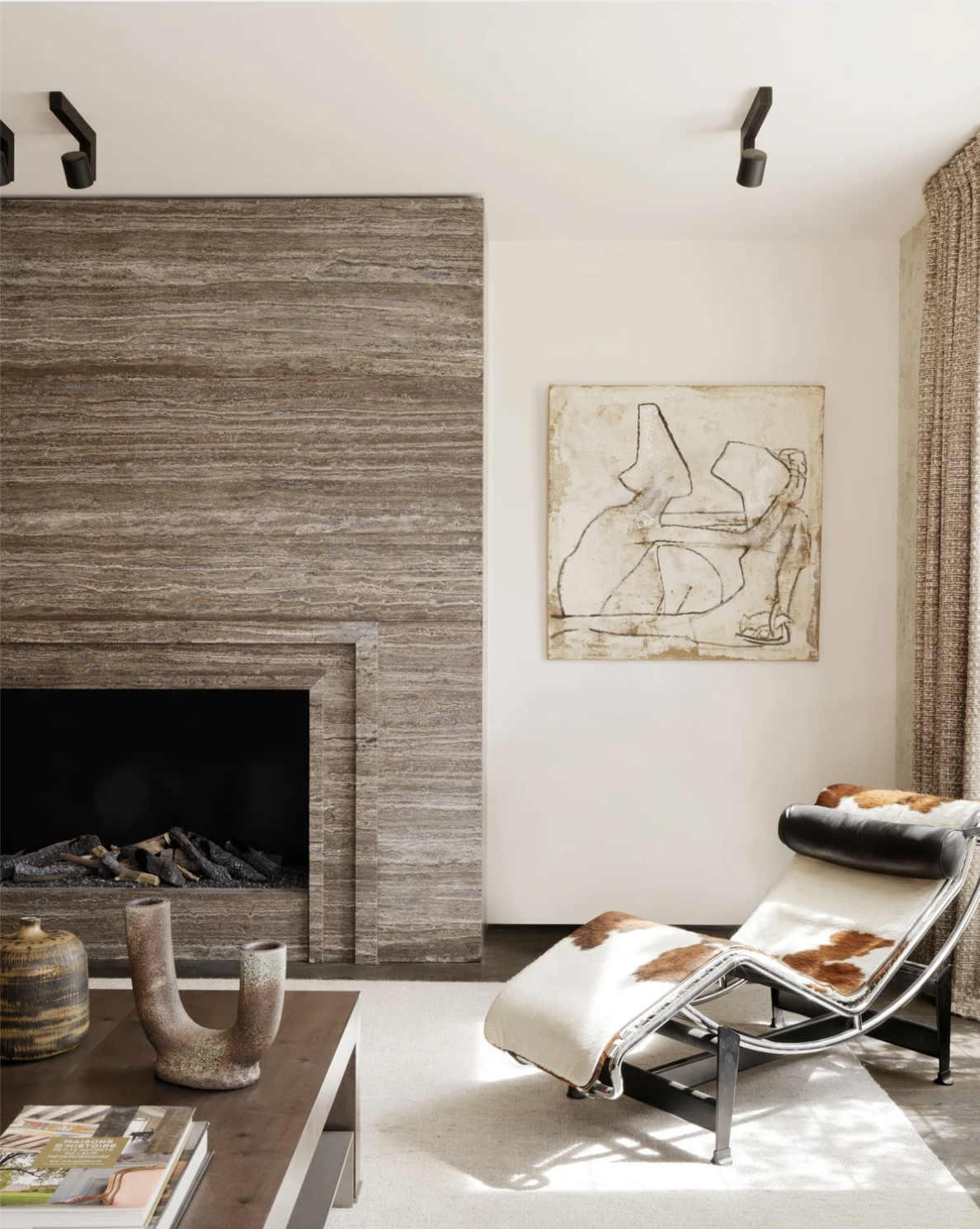 Salon moderne avec cheminée en pierre, fauteuil en cuir et peau de vache, tableau abstrait et décoration artisanale, ambiance chaleureuse et élégante.