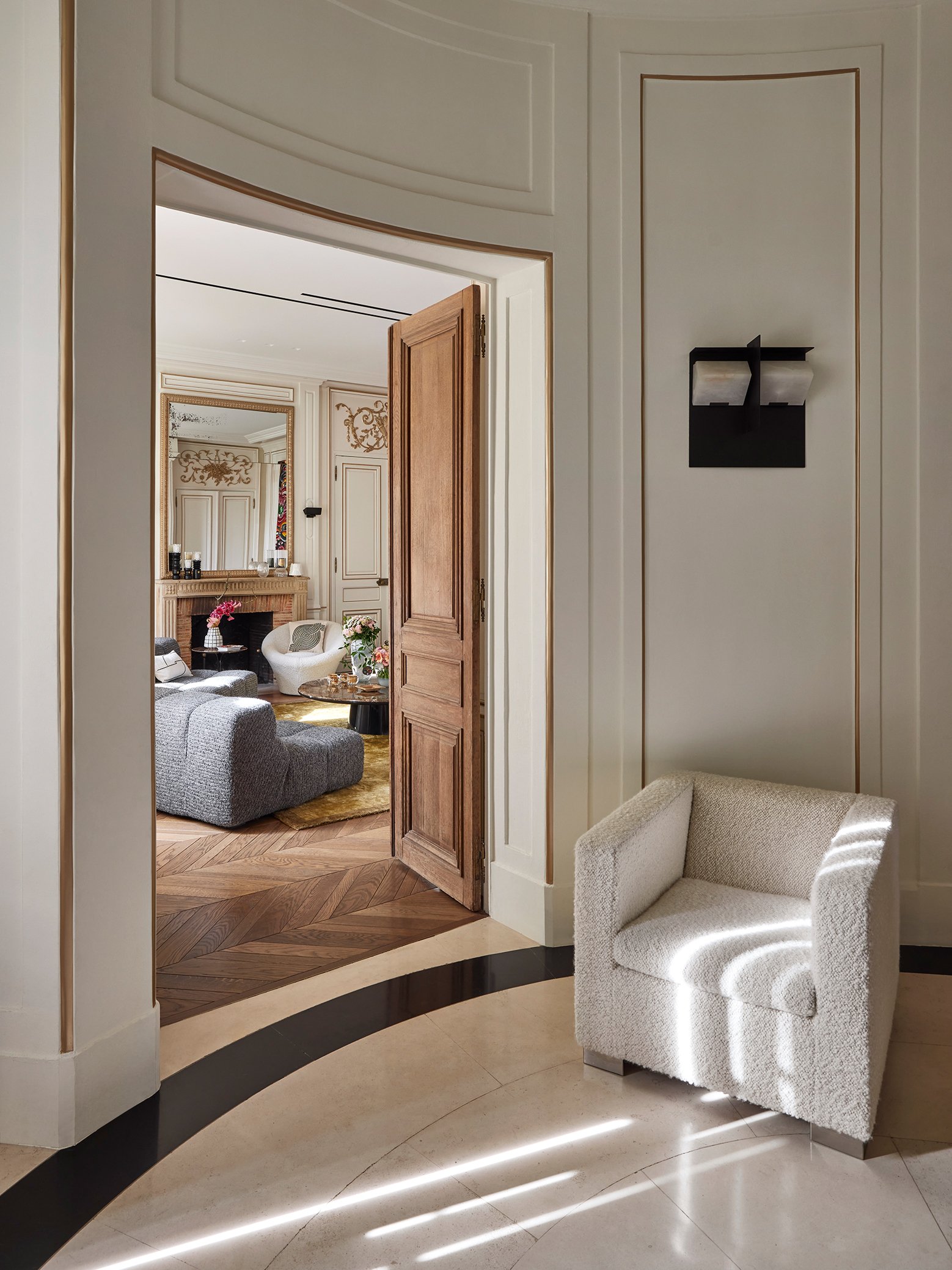 Vue d'un intérieur avec un fauteuil blanc dans le coin droit, une porte en bois ouverte menant à un salon décoré avec un canapé gris, un miroir au-dessus de la cheminée, et quelques objets décoratifs.